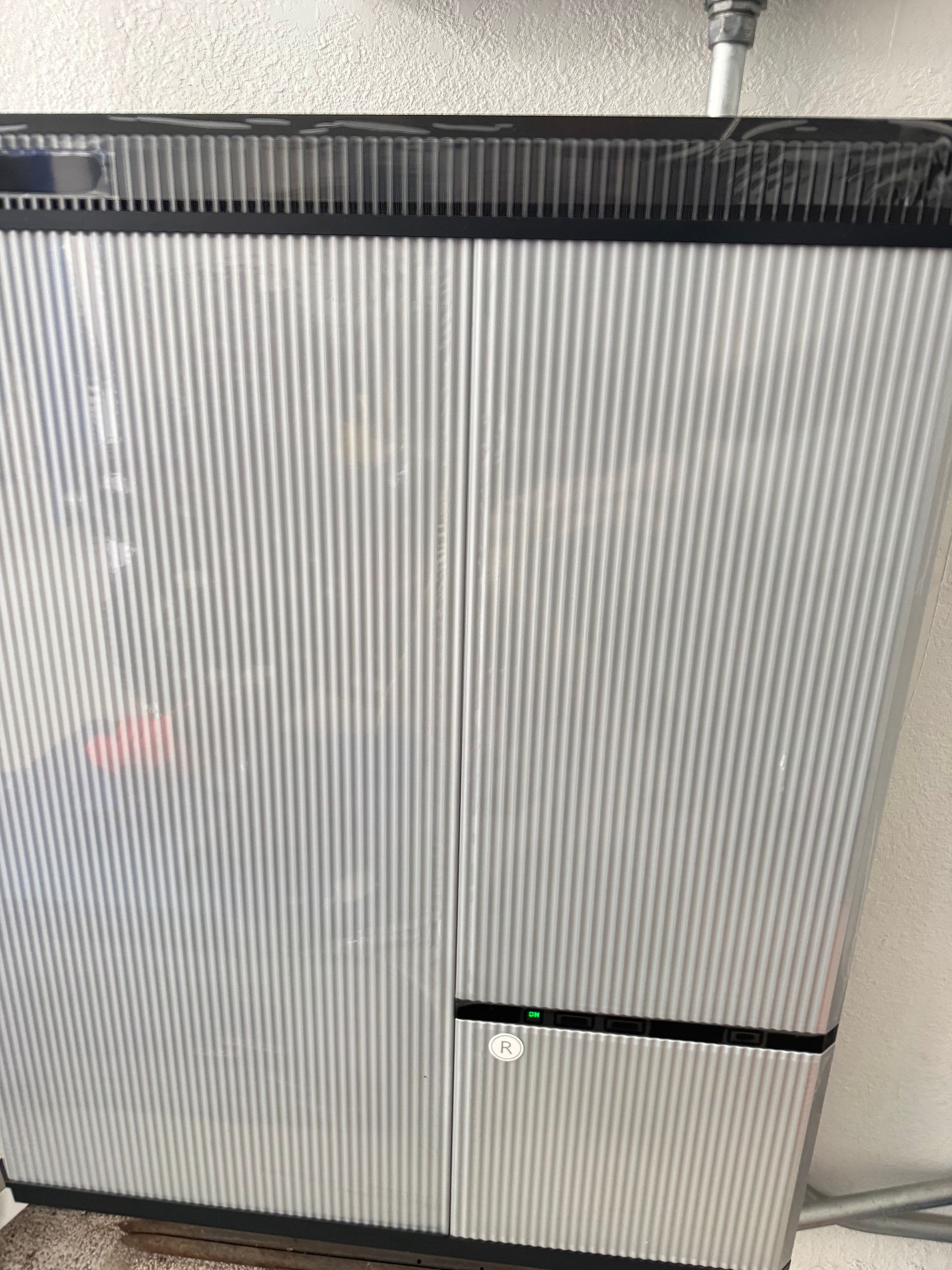 A wall-mounted residential solar battery unit with a metallic, ribbed, silver-grey exterior and a small green light.