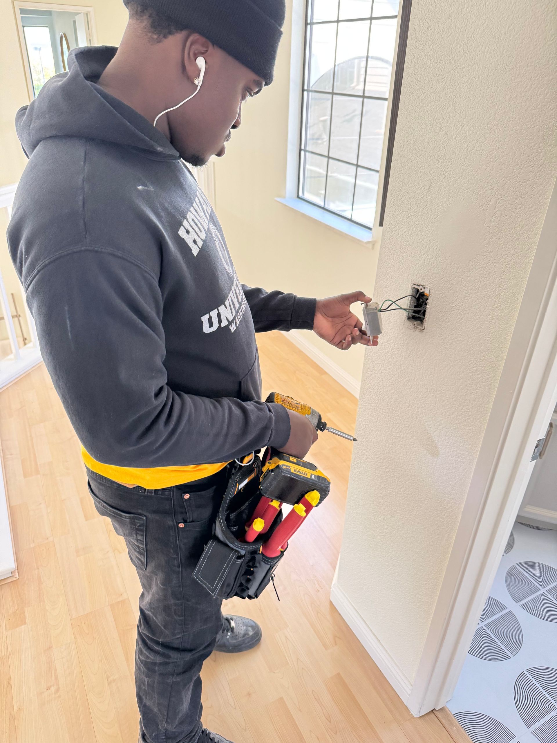 A person wearing a grey hoodie and a tool belt uses a drill to work on electrical wiring in a wall box indoors.