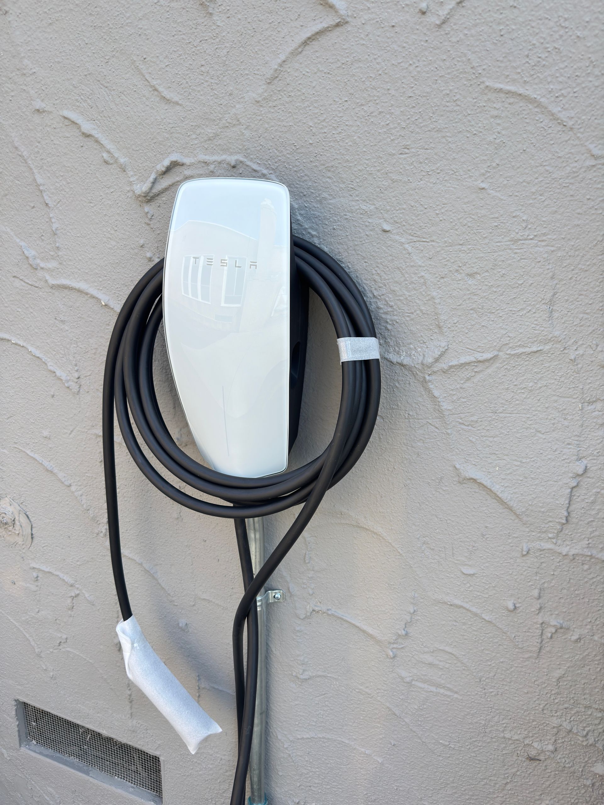 A white electric vehicle charging station mounted on a textured beige wall, with a coiled black charging cable.