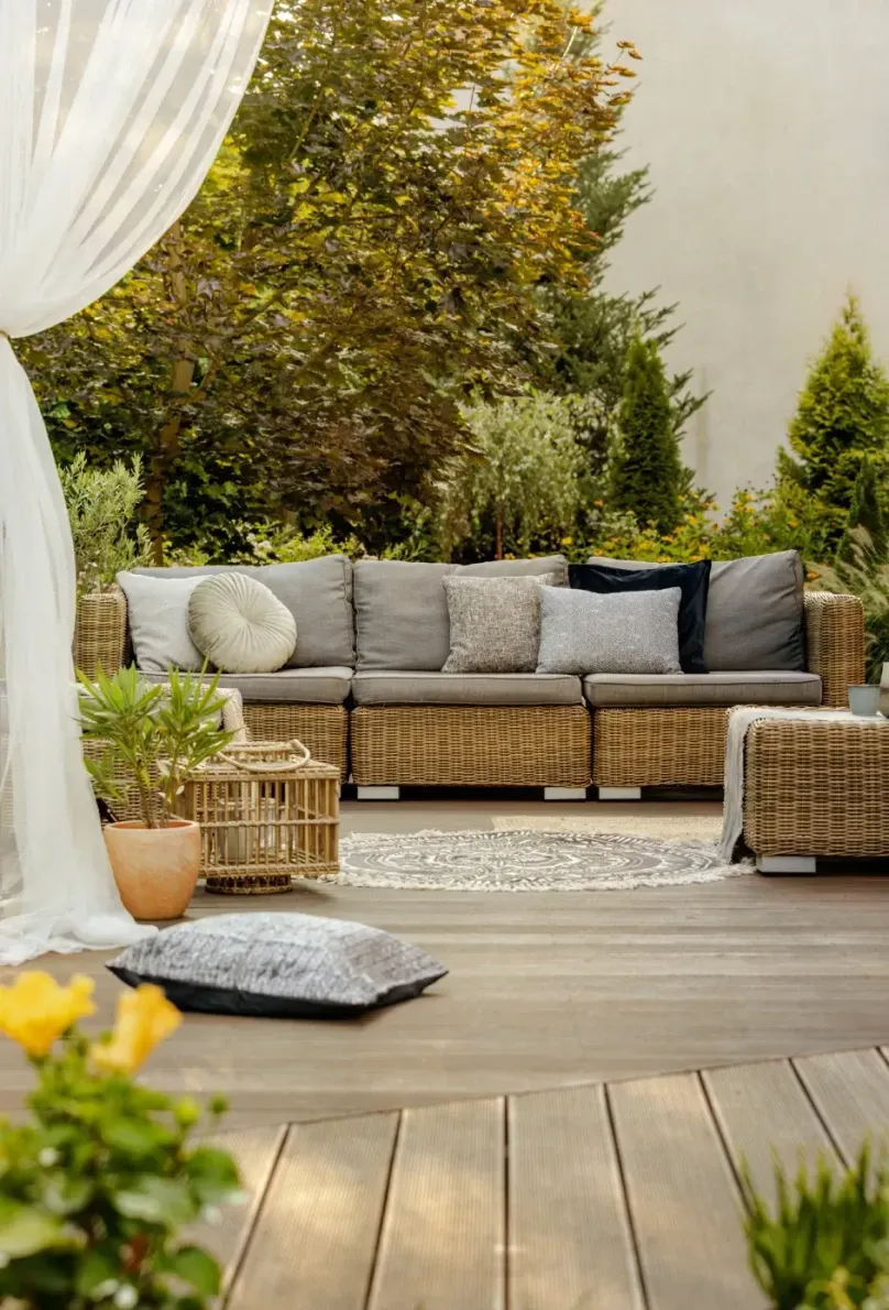 Cozy outdoor patio with woven sofa, cushions, potted plants, and wooden deck in warm sunlight