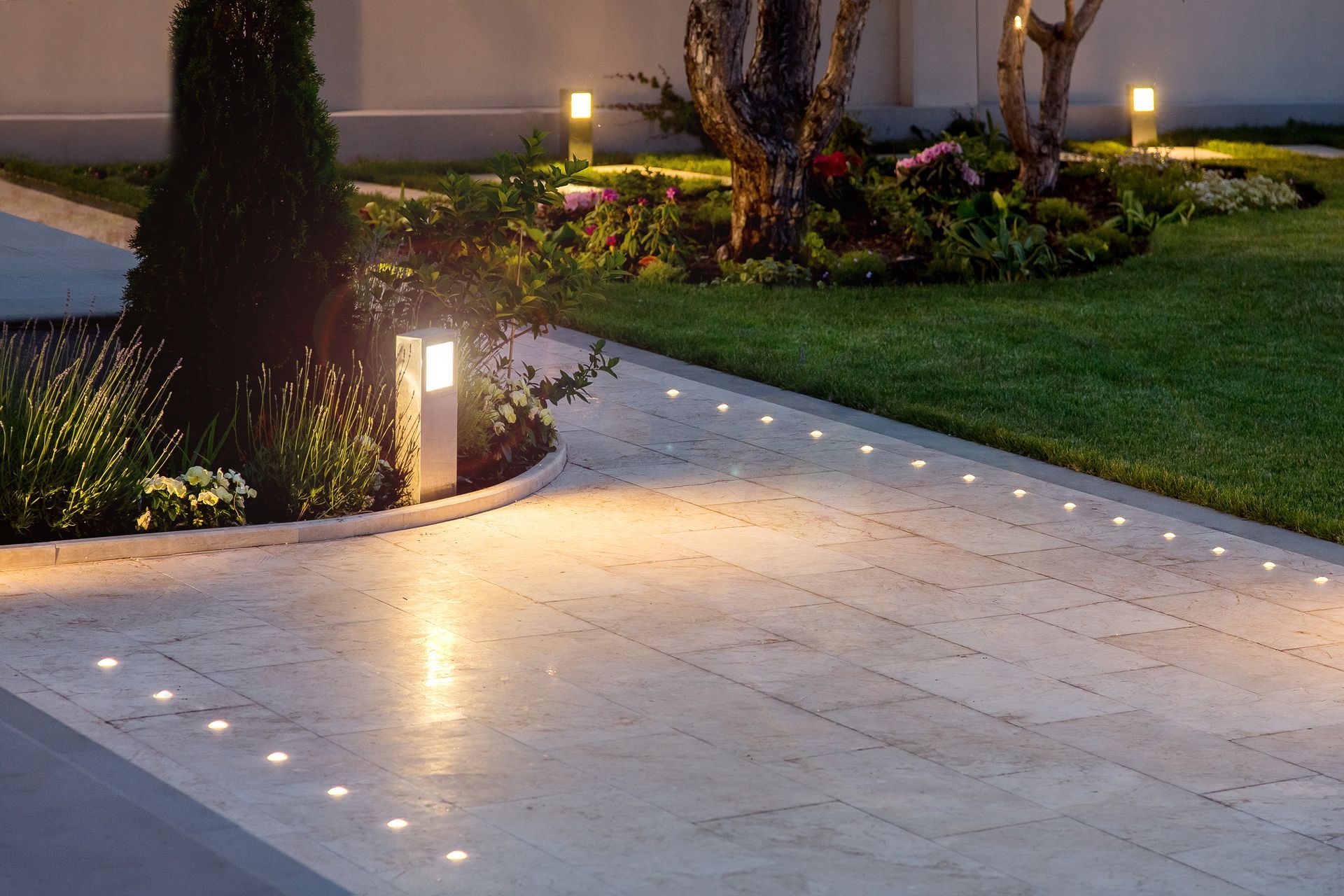 Lit walkway beside a landscaped garden at dusk, with glowing path lights and trees.