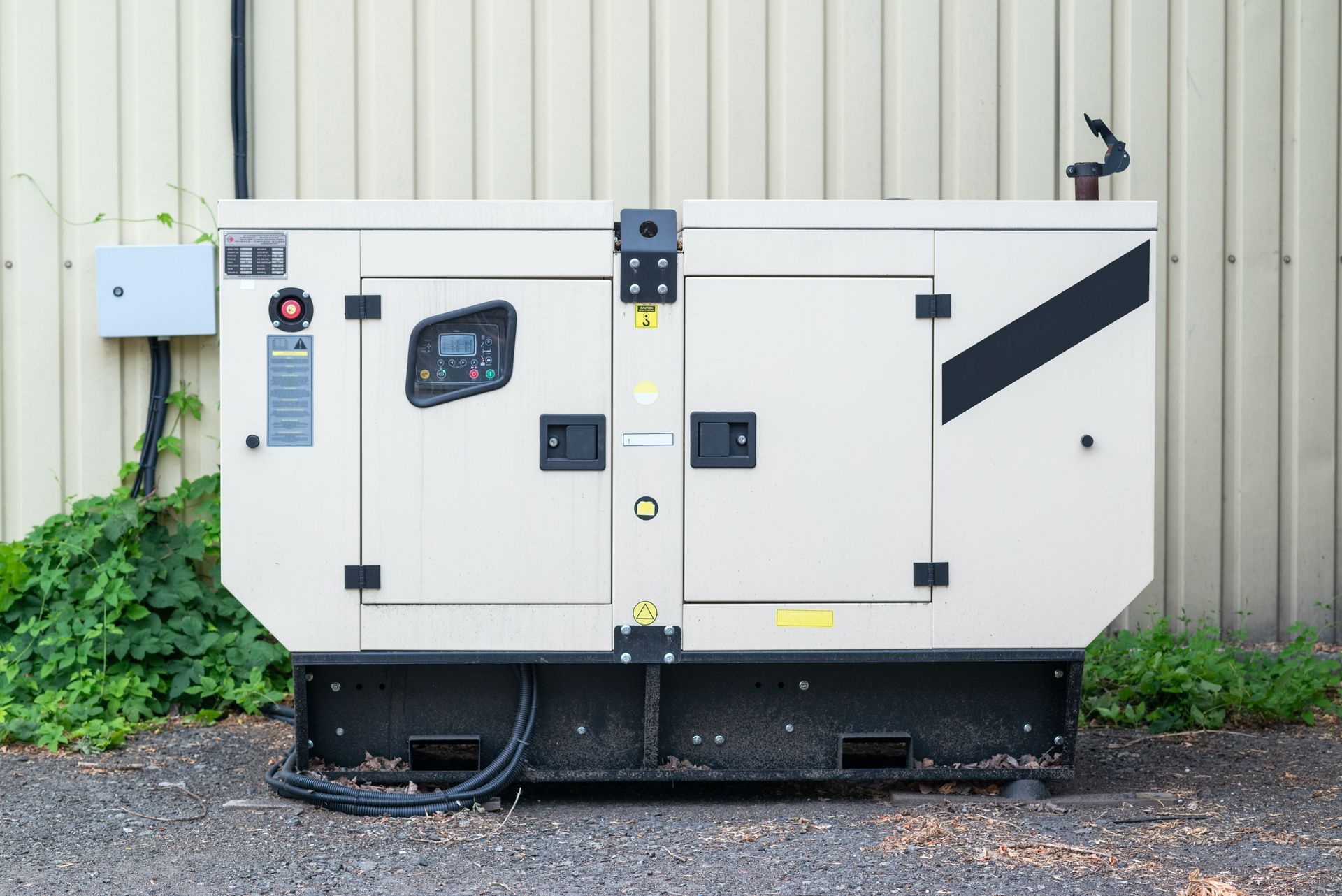 White generator unit beside a corrugated metal wall, with green vines and gravel ground.
