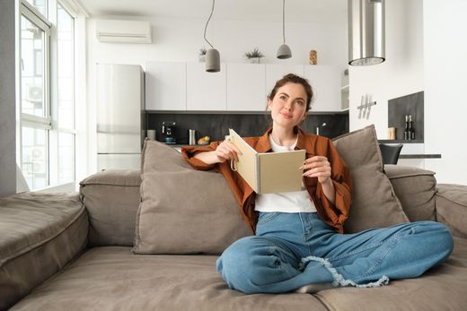 Person sitting cross-legged on a sofa, holding an open notebook in a bright living room