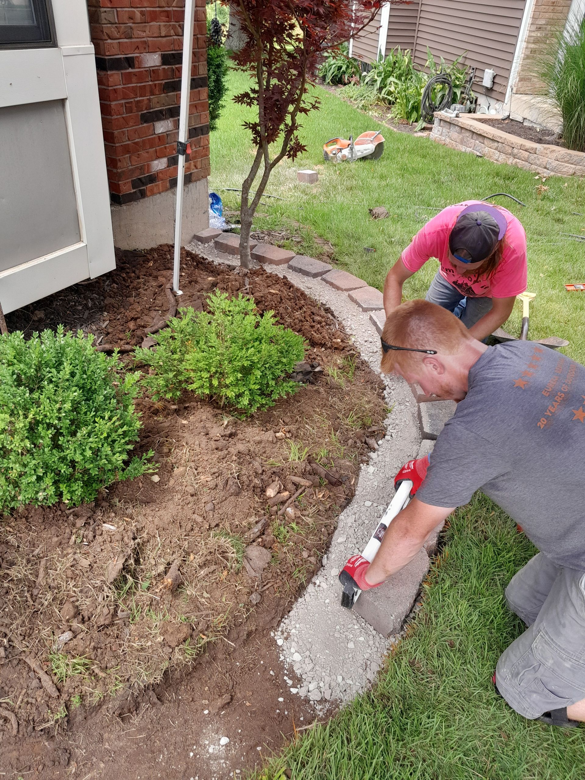 Two people installing a stone border in a garden bed. One kneels applying sealant, the other works behind them.
