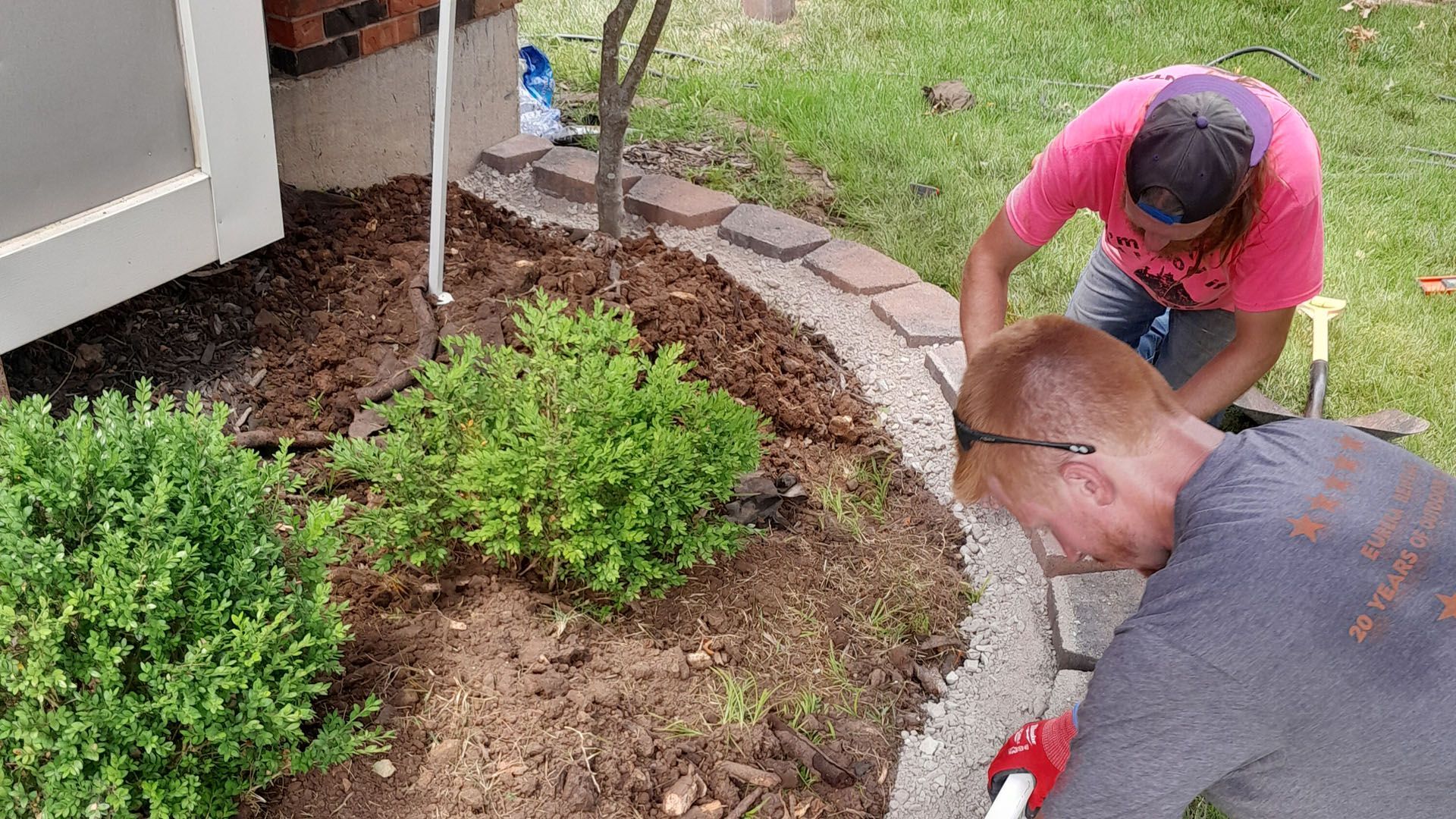 Two people installing a brick border around a garden bed. One kneels, the other bends over; both are working with bricks.