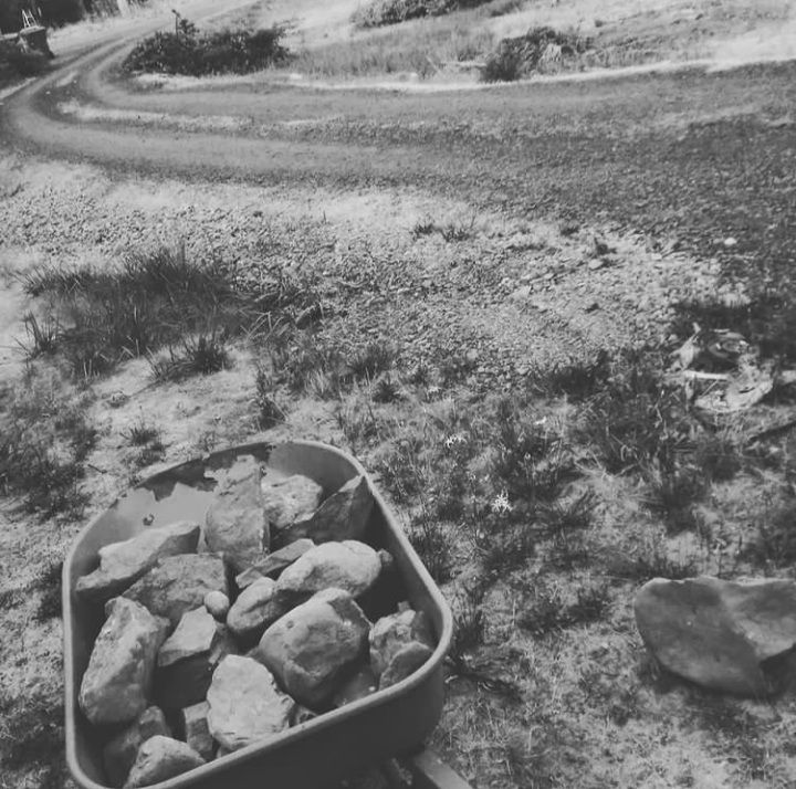 Wheelbarrow filled with rocks on a dirt path, with a winding road in the background.