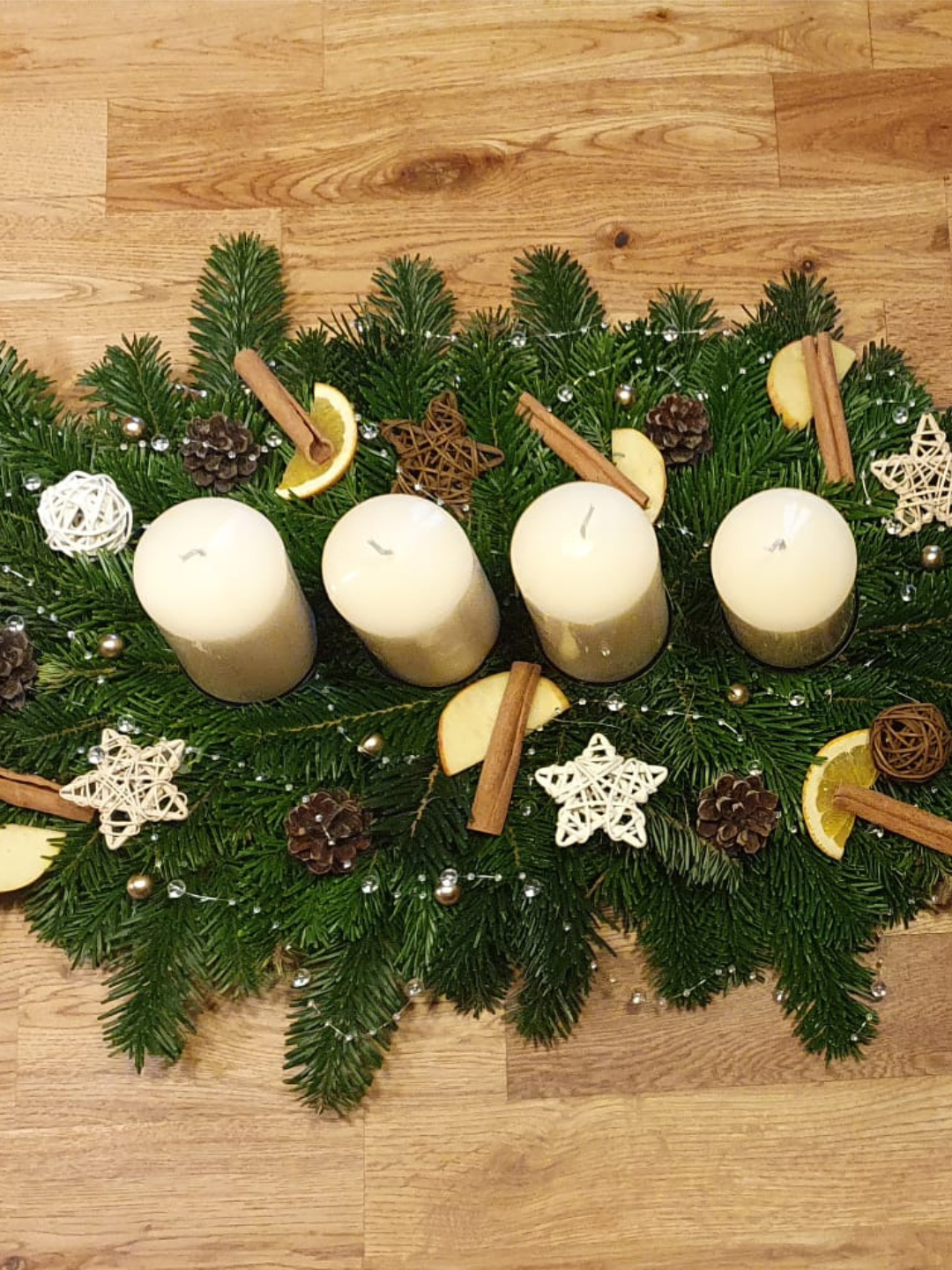 Quatre bougies blanches sur une couronne de sapin ornée de pommes de pin, d'étoiles en bois et de bâtonnets de cannelle.
