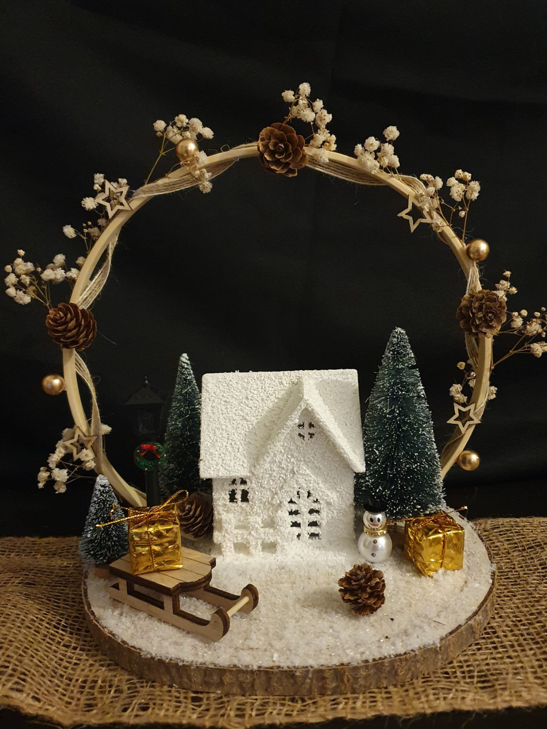 Petite maison enneigée avec des pins, un traîneau et une couronne décorée de pommes de pin sur une table rustique