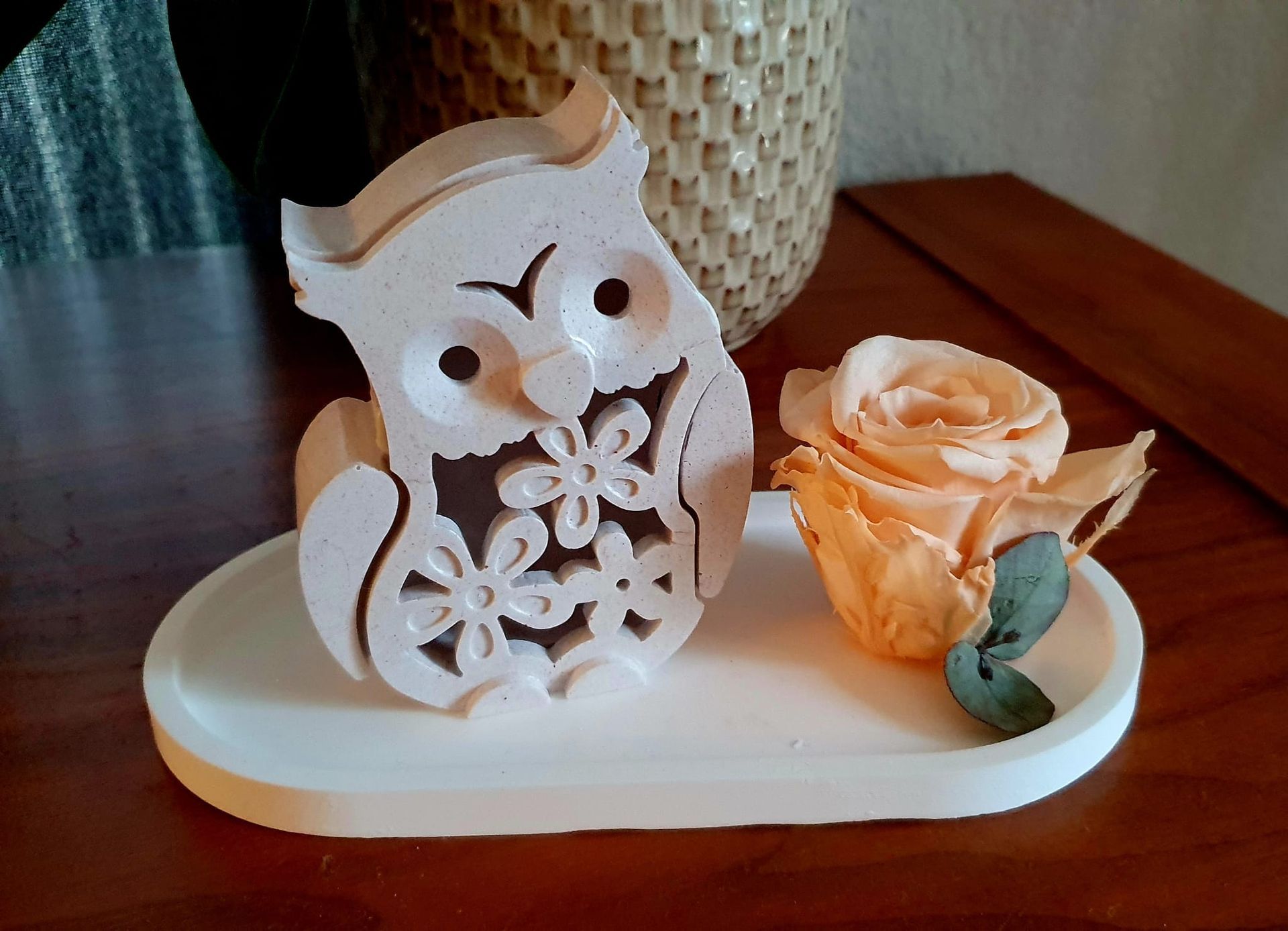 Figurine de hibou blanc à côté d'une rose pêche sur un plateau blanc, avec un vase tressé en arrière-plan.