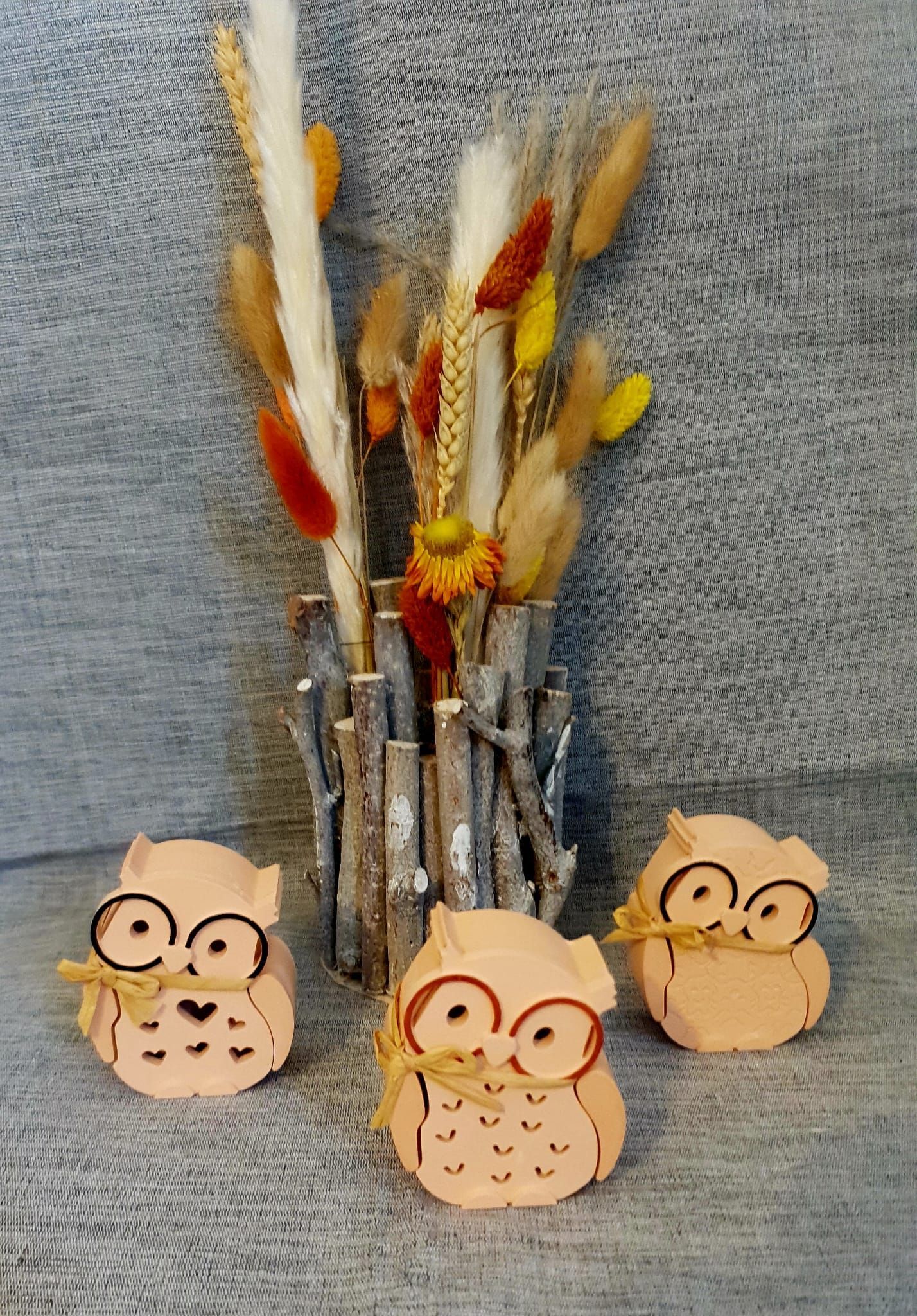 Trois ornements en forme de tête de hibou avec des lunettes rouges dans un vase contenant de hautes tiges séchées.