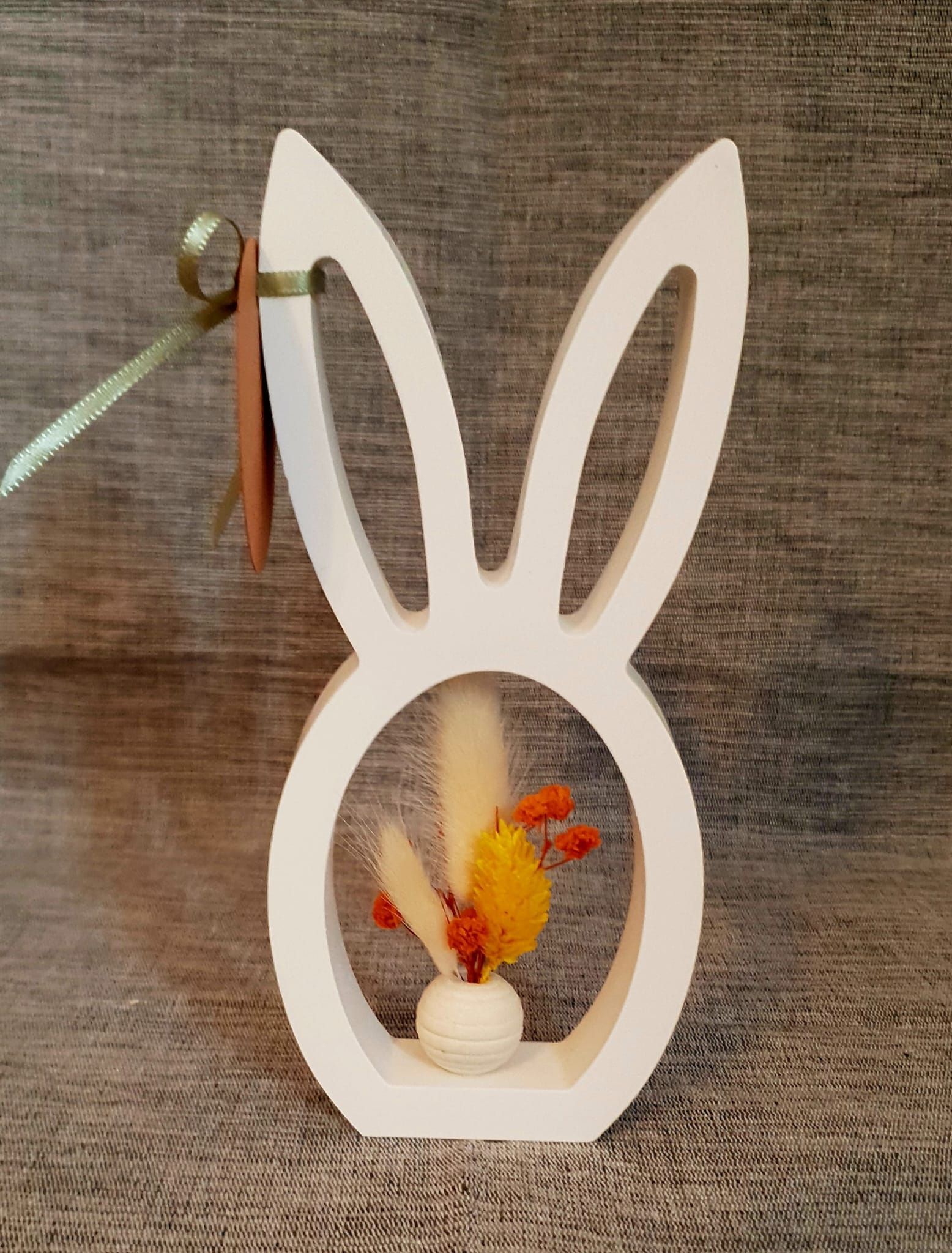 Décoration en forme de lapin blanc avec des fleurs séchées sur un fond gris texturé.