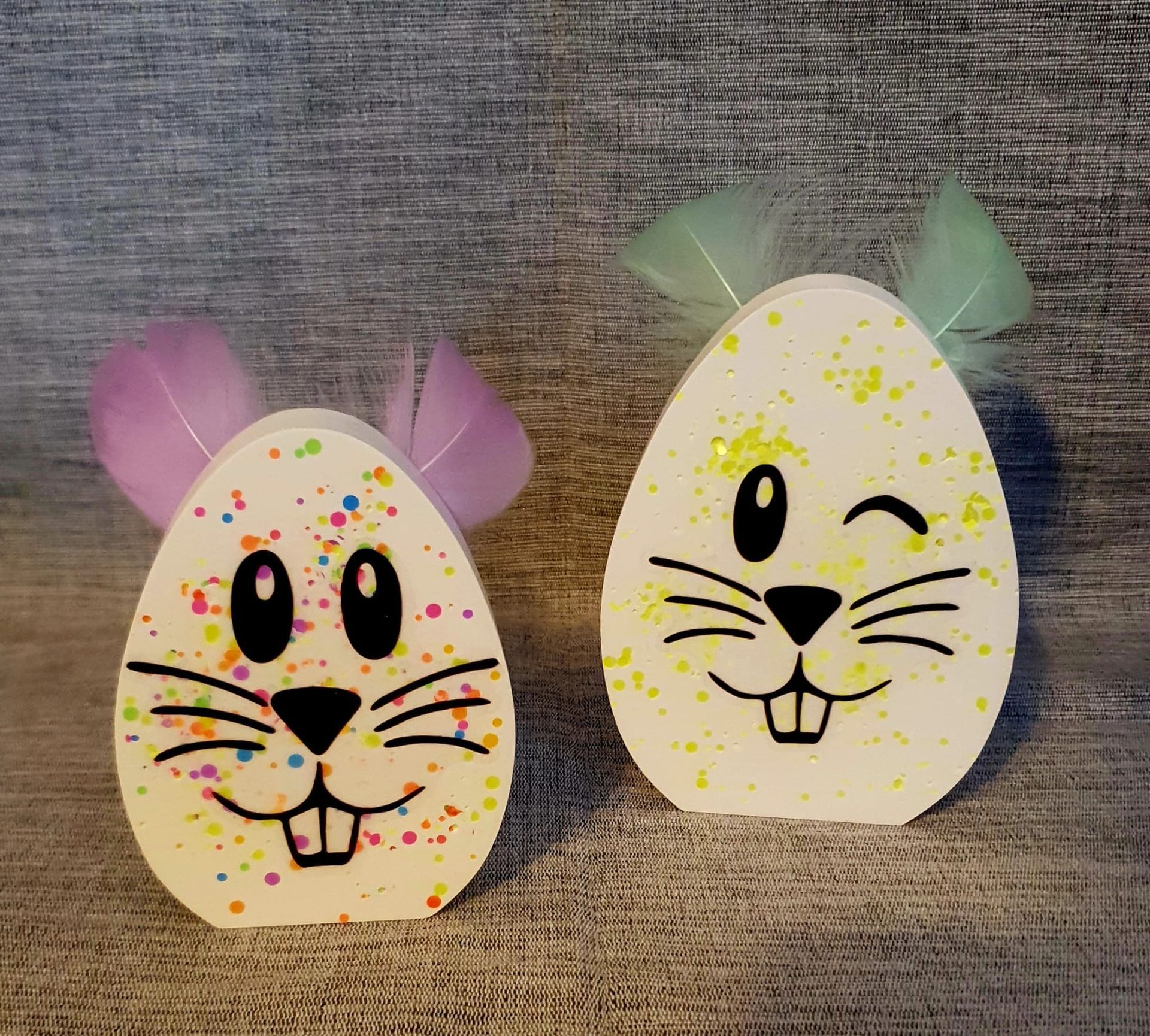 Deux têtes de lapin décorées en forme d'œuf sur un fond de tissu gris, l'une faisant un clin d'œil, l'autre souriant