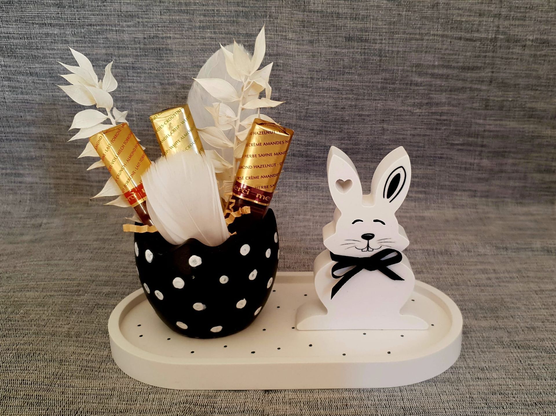 Vase noir à pois orné de fleurs blanches et dorées, posé à côté d'une petite figurine de lapin blanc sur un plateau.