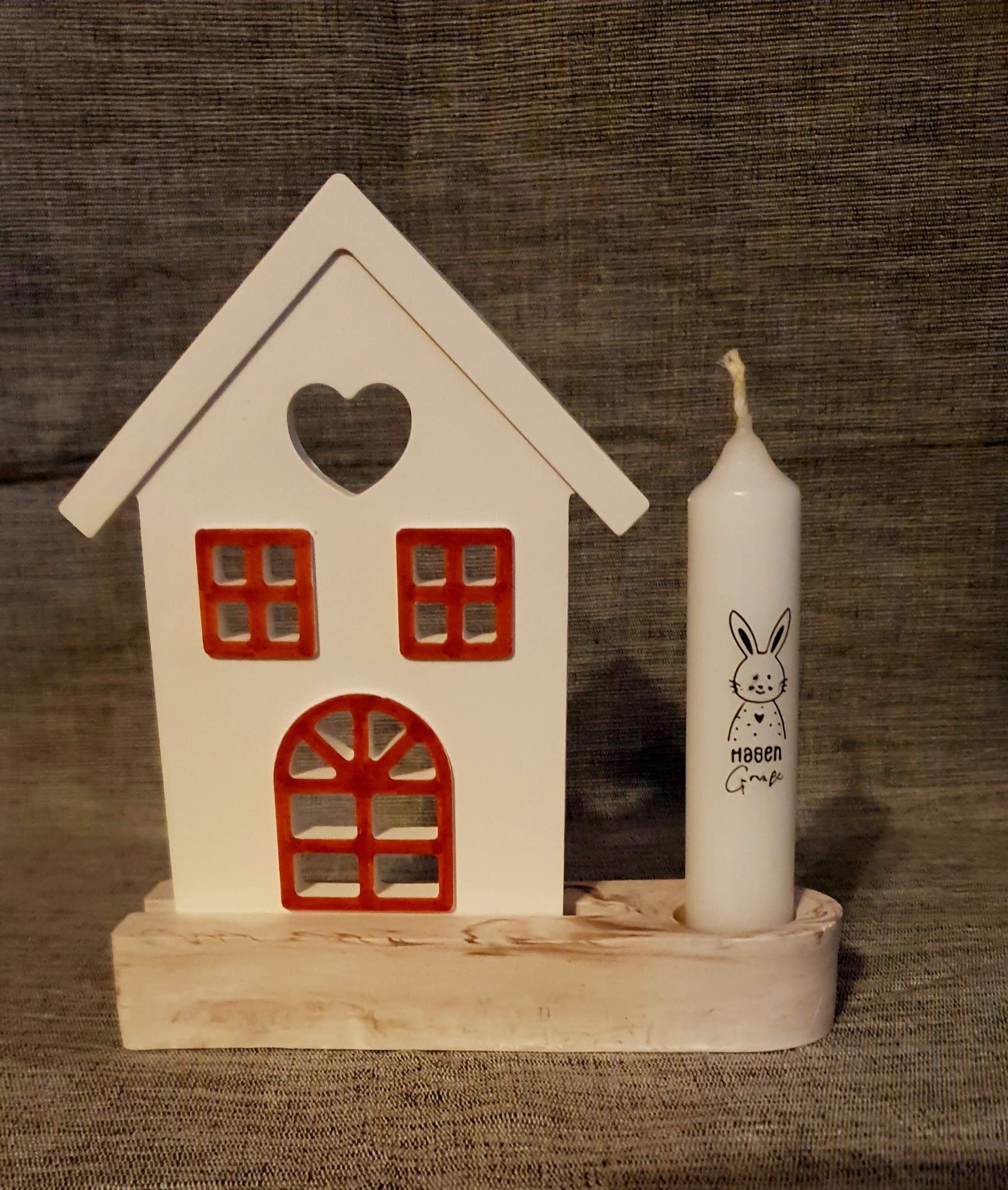 Figurine de maison en bois blanc avec fenêtres rouges et découpe en forme de cœur, posée à côté d'une bougie blanche.