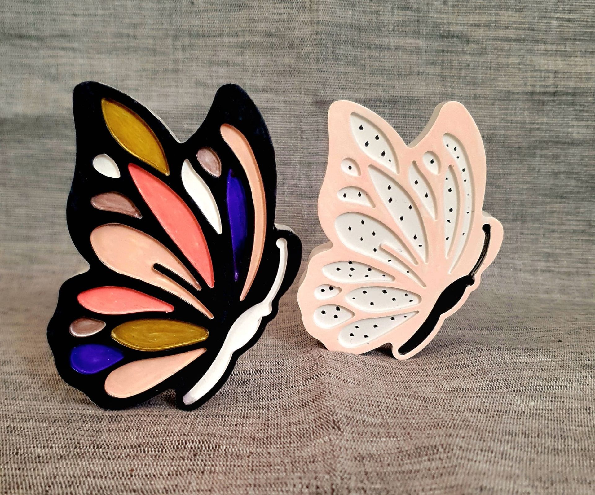 Deux ornements décoratifs en forme de papillon, l'un noir aux ailes colorées et l'autre rose pâle, sur une surface grise