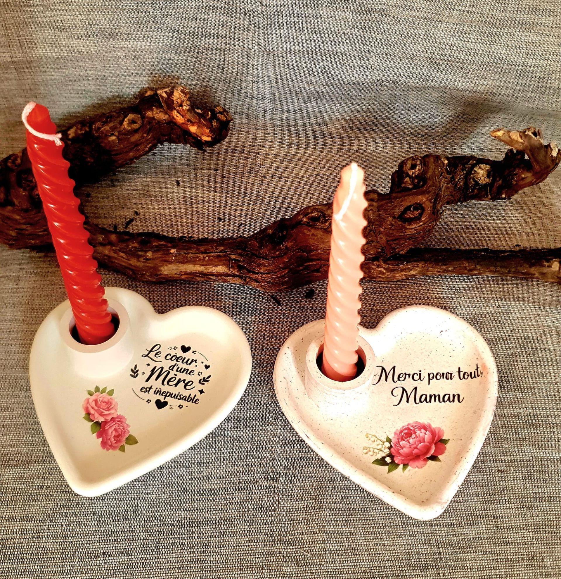 Deux bougeoirs en forme de cœur avec des bougies rouges et roses sur du bois, l'un portant l'inscription « Merci maman ».