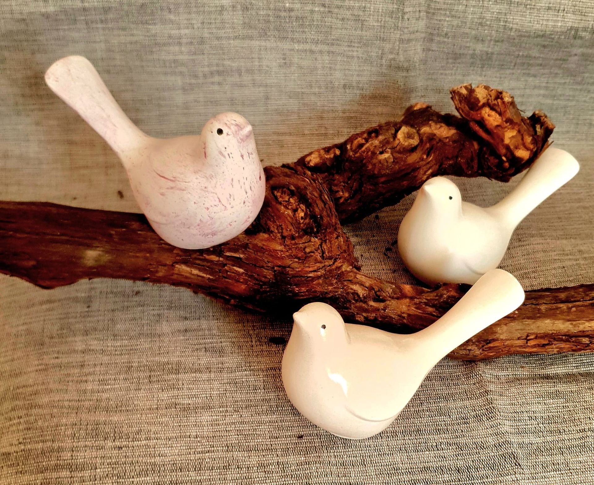 Trois figurines d'oiseaux blancs sur une branche de bois rustique, sur fond de toile de jute.