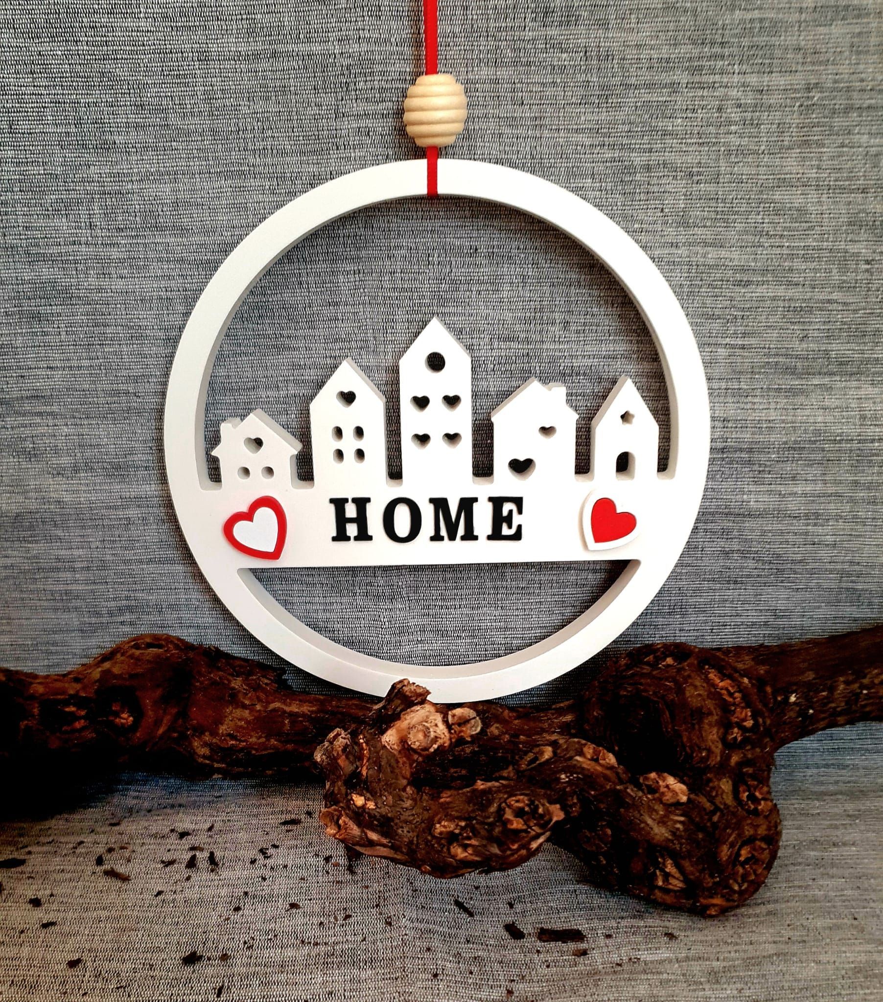 Ornement circulaire blanc HOME orné de cœurs rouges et d'une petite silhouette de village, suspendu à une ficelle rouge.