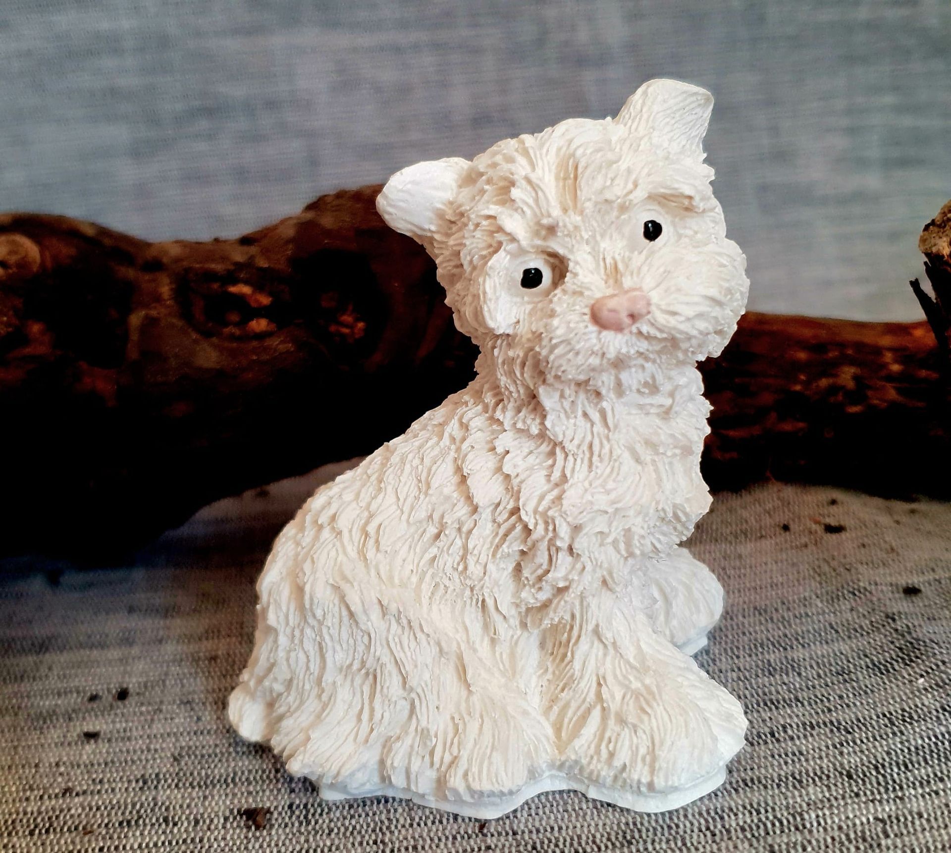 Petite figurine de chat en céramique blanche, au pelage texturé et au nez rose, posée sur un support en tissu.
