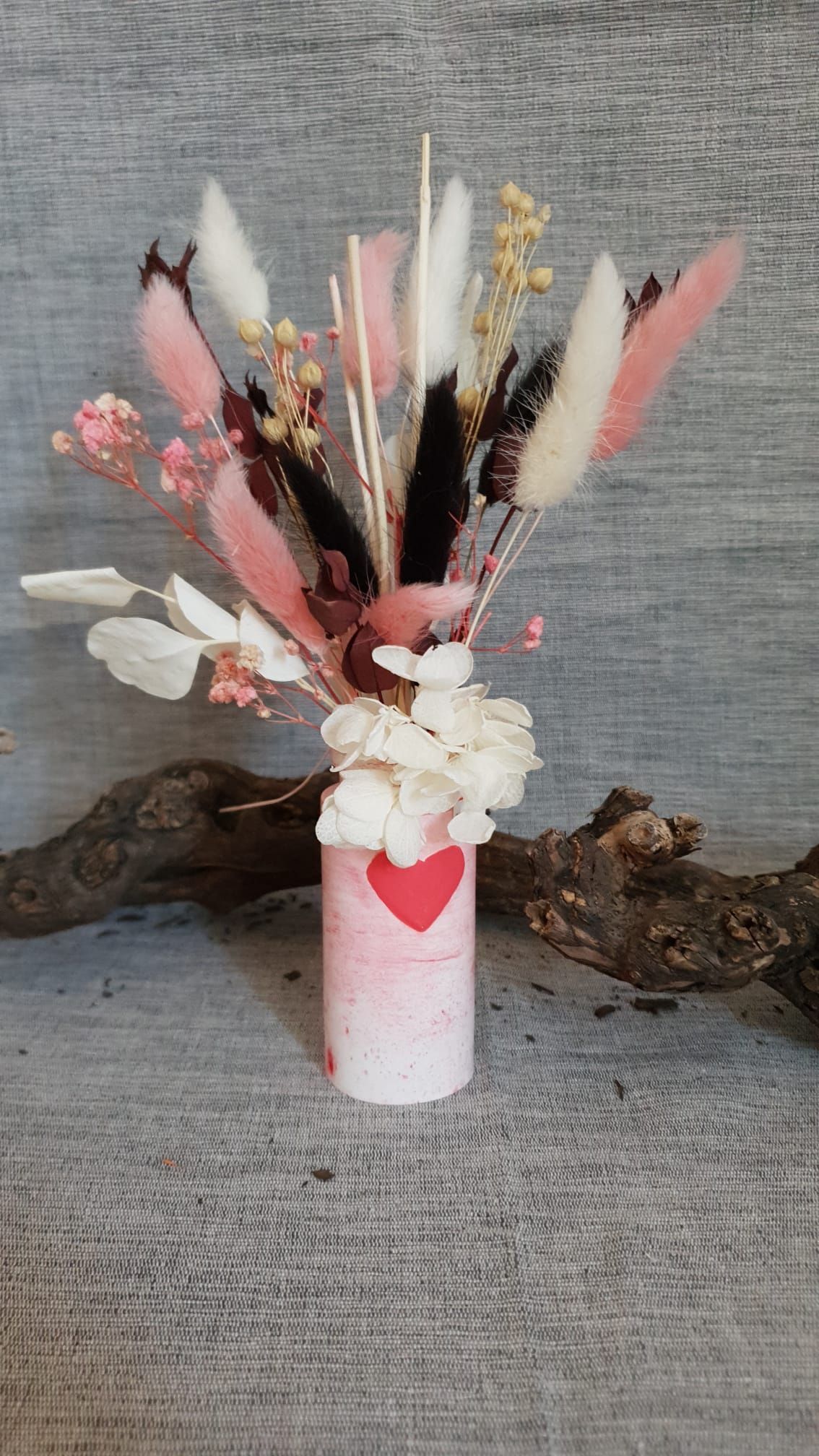 Vase blanc orné d'un cœur rouge contenant un arrangement de fleurs séchées sur fond de tissu gris