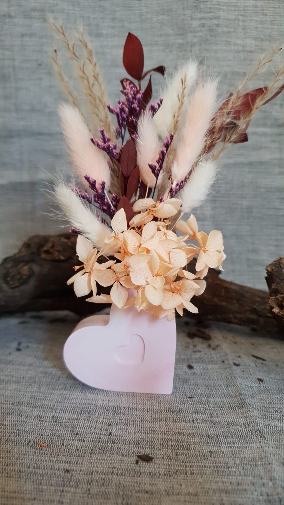 Petit arrangement de fleurs séchées dans un support rose en forme de cœur, posé sur une surface rustique.