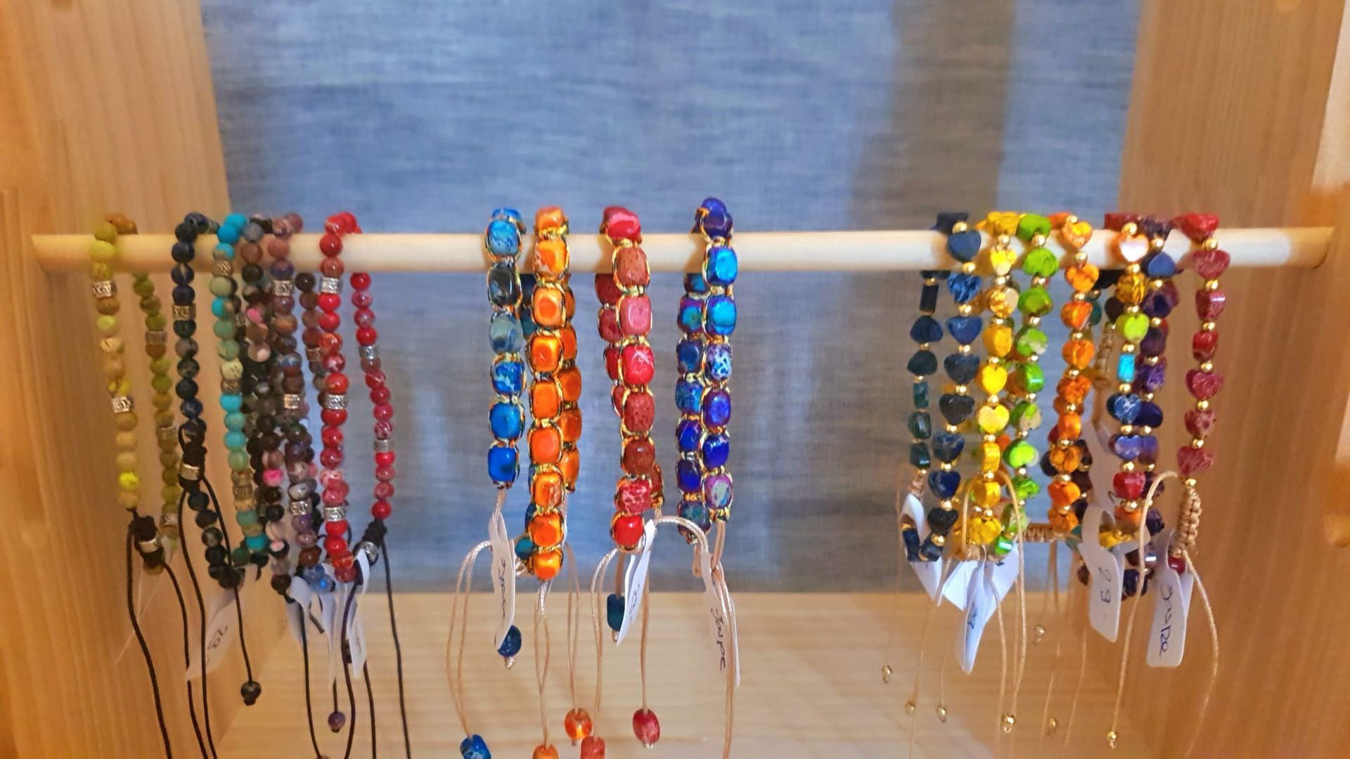 Des colliers de perles colorées pendent d'une tringle blanche, soigneusement disposés en rangées sur un mur beige.