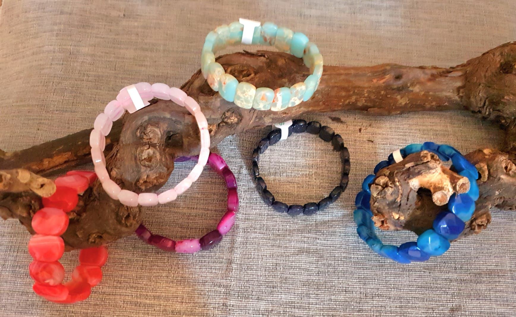 Cinq bracelets de perles colorées disposés sur un morceau de bois flotté, lui-même posé sur une surface tissée.