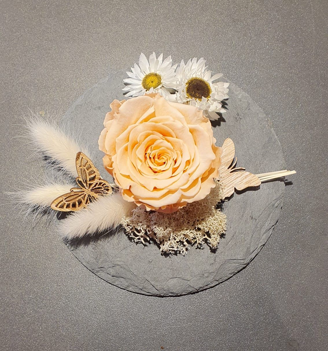 Centre de table composé de roses pêche, de marguerites blanches et d'herbes séchées sur une surface grise