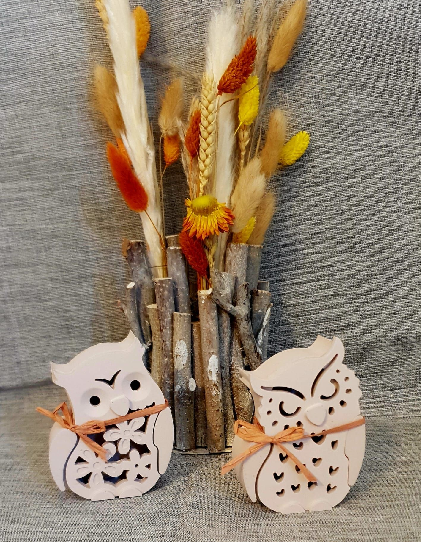 Deux figurines de hibou à côté de fleurs séchées dans un vase rustique sur un fond de tissu gris