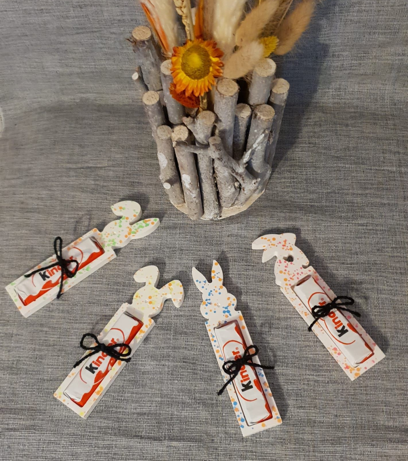 Surface grise texturée avec un vase de fleurs séchées et quatre petits cadeaux artisanaux noués disposés en dessous.
