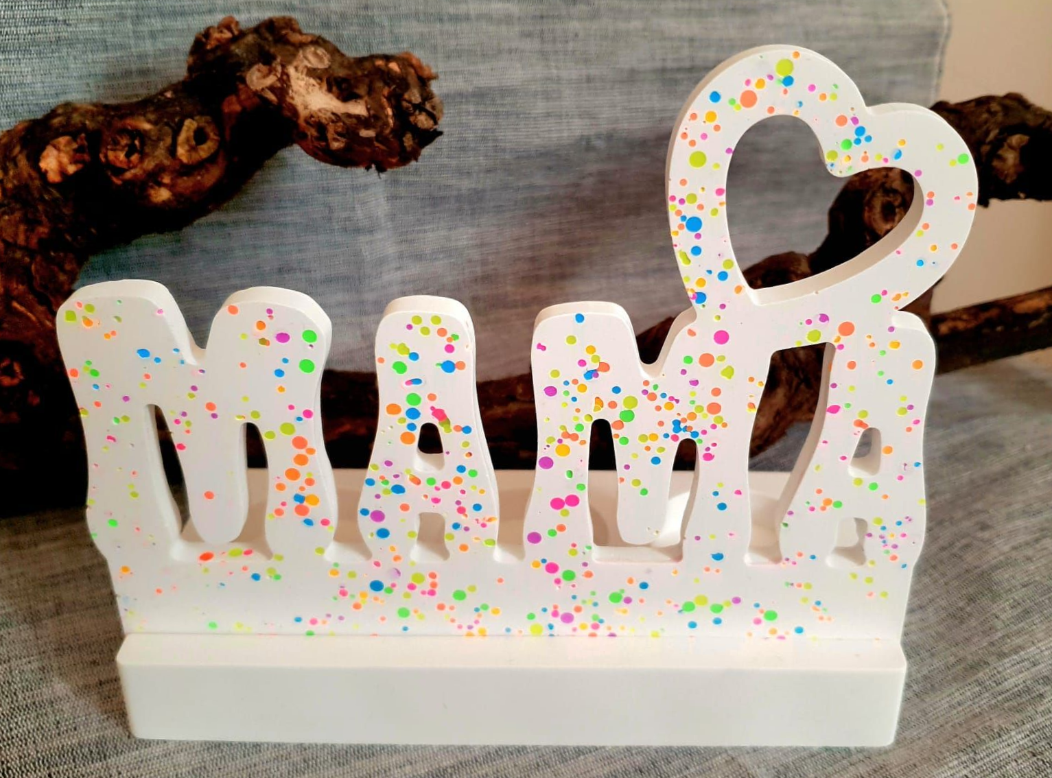 Une pancarte blanche pailletée « MAMA » ornée de confettis colorés est posée sur une étagère avec une branche décorative.