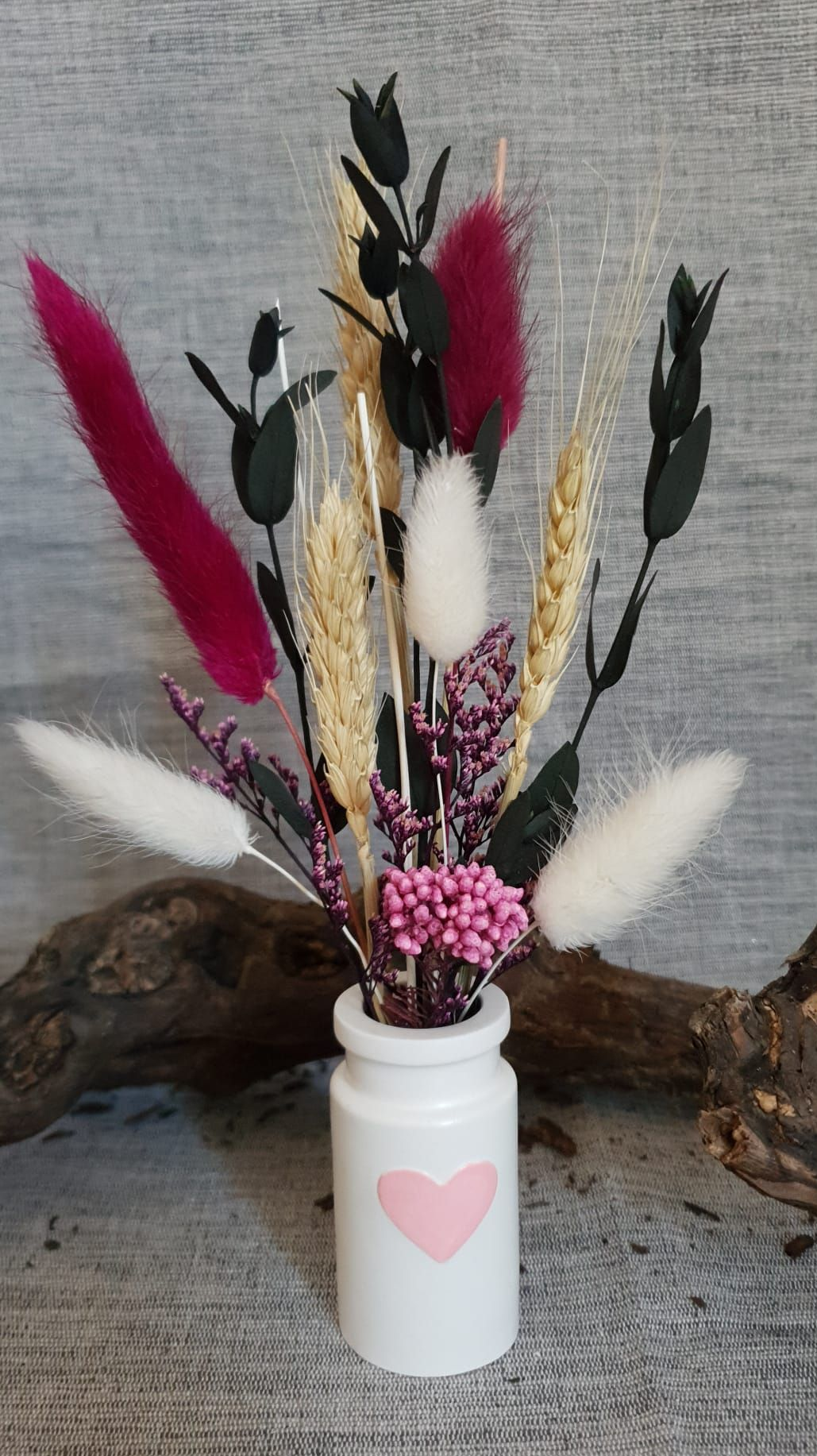 Composition florale séchée décorative dans un vase blanc avec un cœur rose, sur une surface en bois