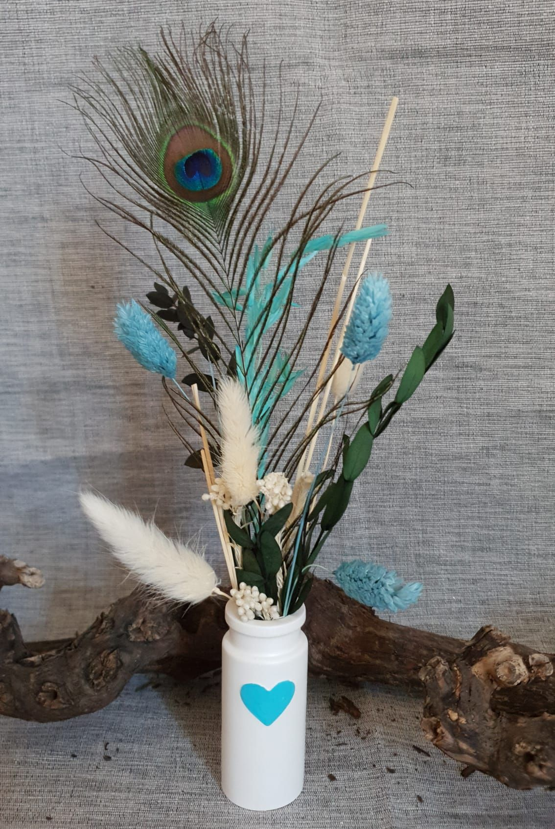 Vase blanc orné d'un cœur bleu contenant des plumes de paon et des fleurs séchées bleu-blanc sur fond de bois flotté
