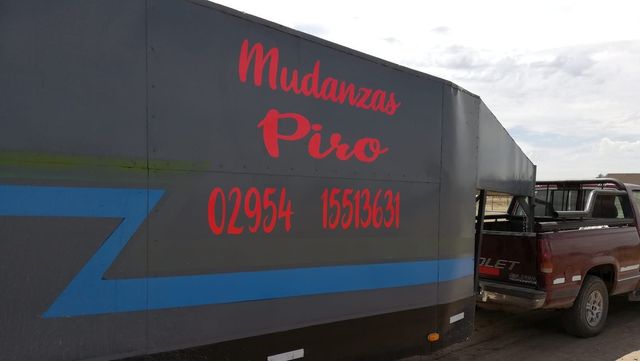 Camión de mudanzas con la inscripción “Mudanzas Piro” en rojo sobre un remolque gris, al lado de una camioneta pickup marrón.