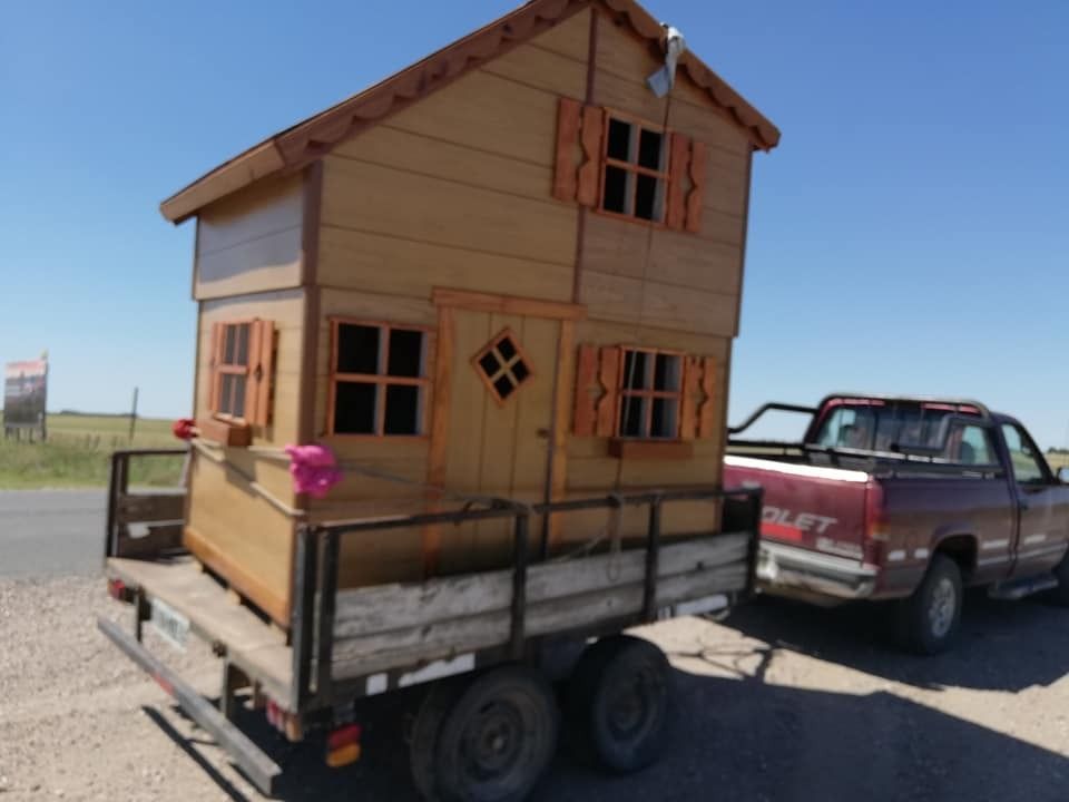 Pequeña casita de juegos de madera sobre un remolque, enganchada a una camioneta roja al borde de la carretera.