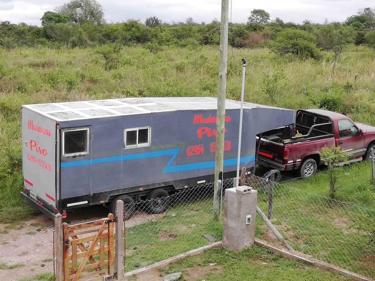 Un camión rojo remolca un remolque gris y azul a través de un campo de hierba cerca de un poste de electricidad.