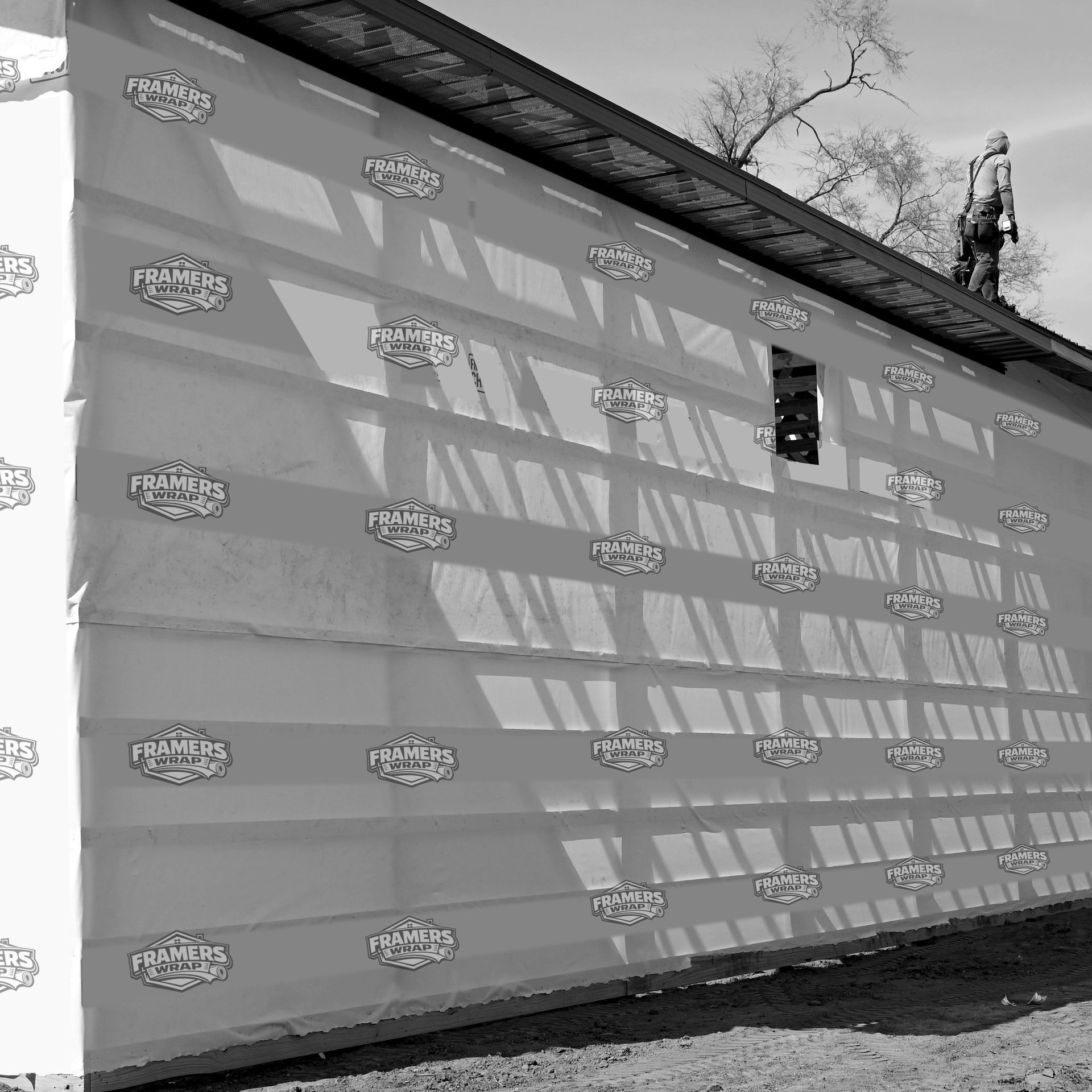 Long white building under construction wrapped in printed protective sheeting, with strong diagonal shadows.