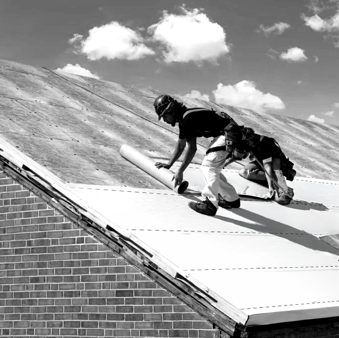 Two workers on a roof installing roofing underlayment under a partly cloudy sky
