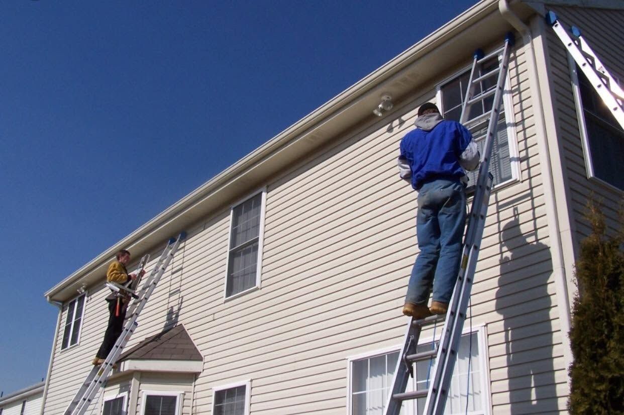 Two workers on ladders painting the side of a white house