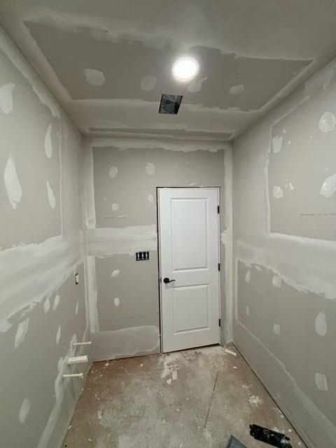 Narrow unfinished drywall room with a white door, ceiling light, and exposed floor.