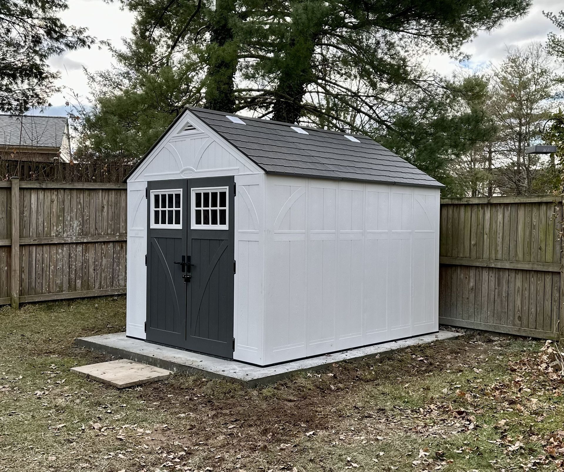 White backyard shed with black double doors and small windows, surrounded by a wooden fence and trees.