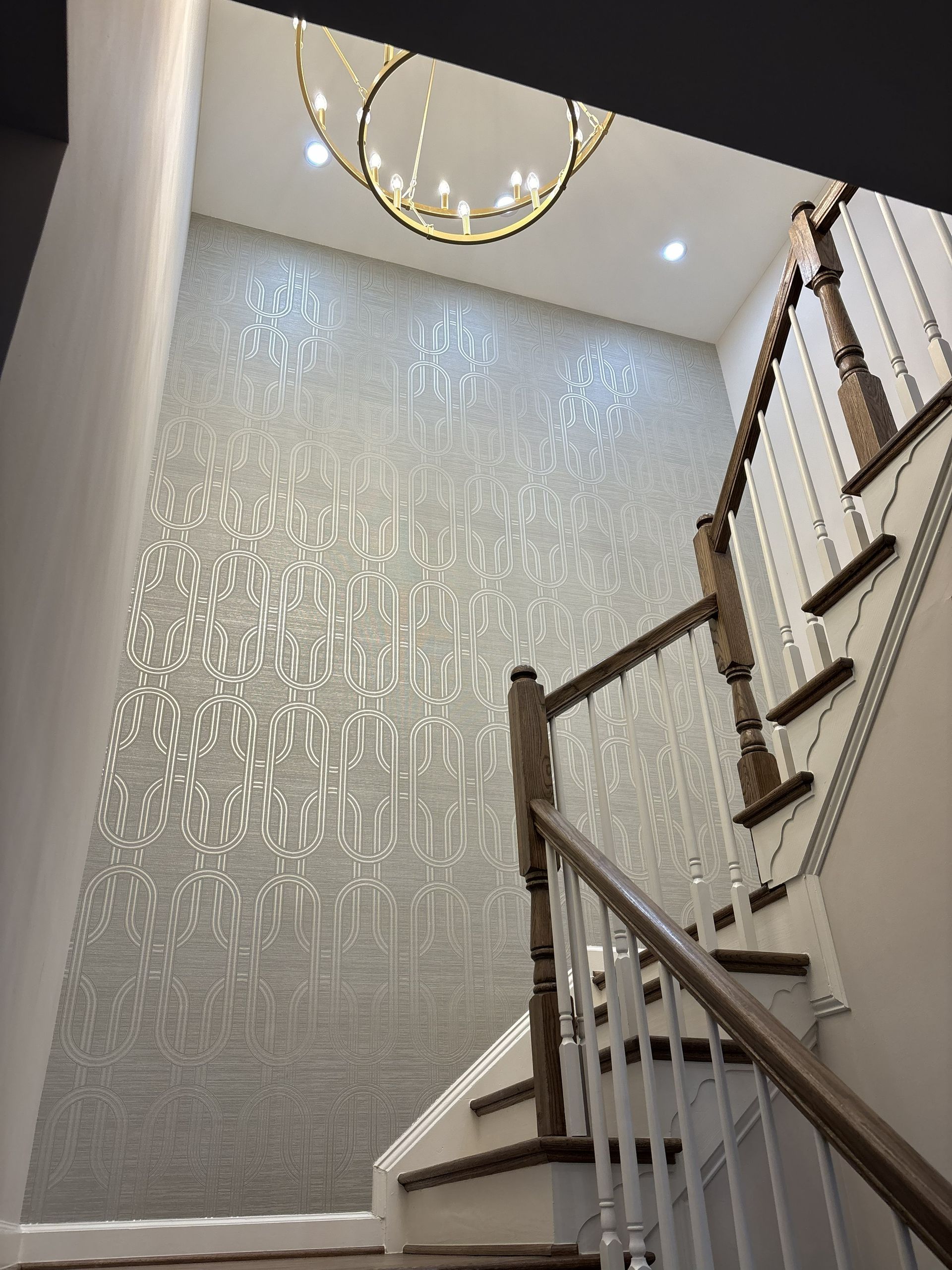 Staircase with wood railings beside a textured gray wall and modern chandelier in a bright foyer