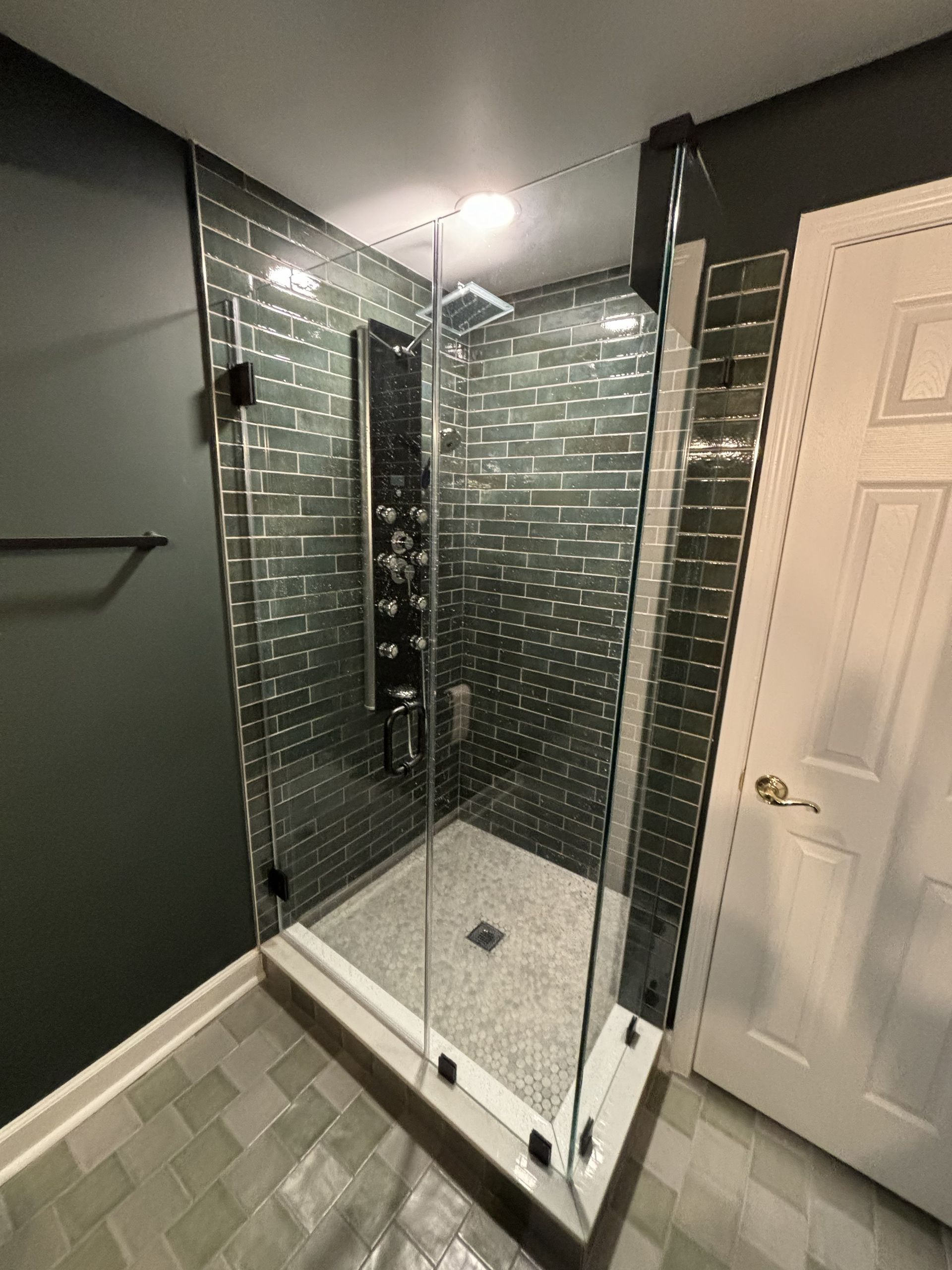 Glass corner shower with chrome frame and gray tiled bathroom walls, next to a white door.