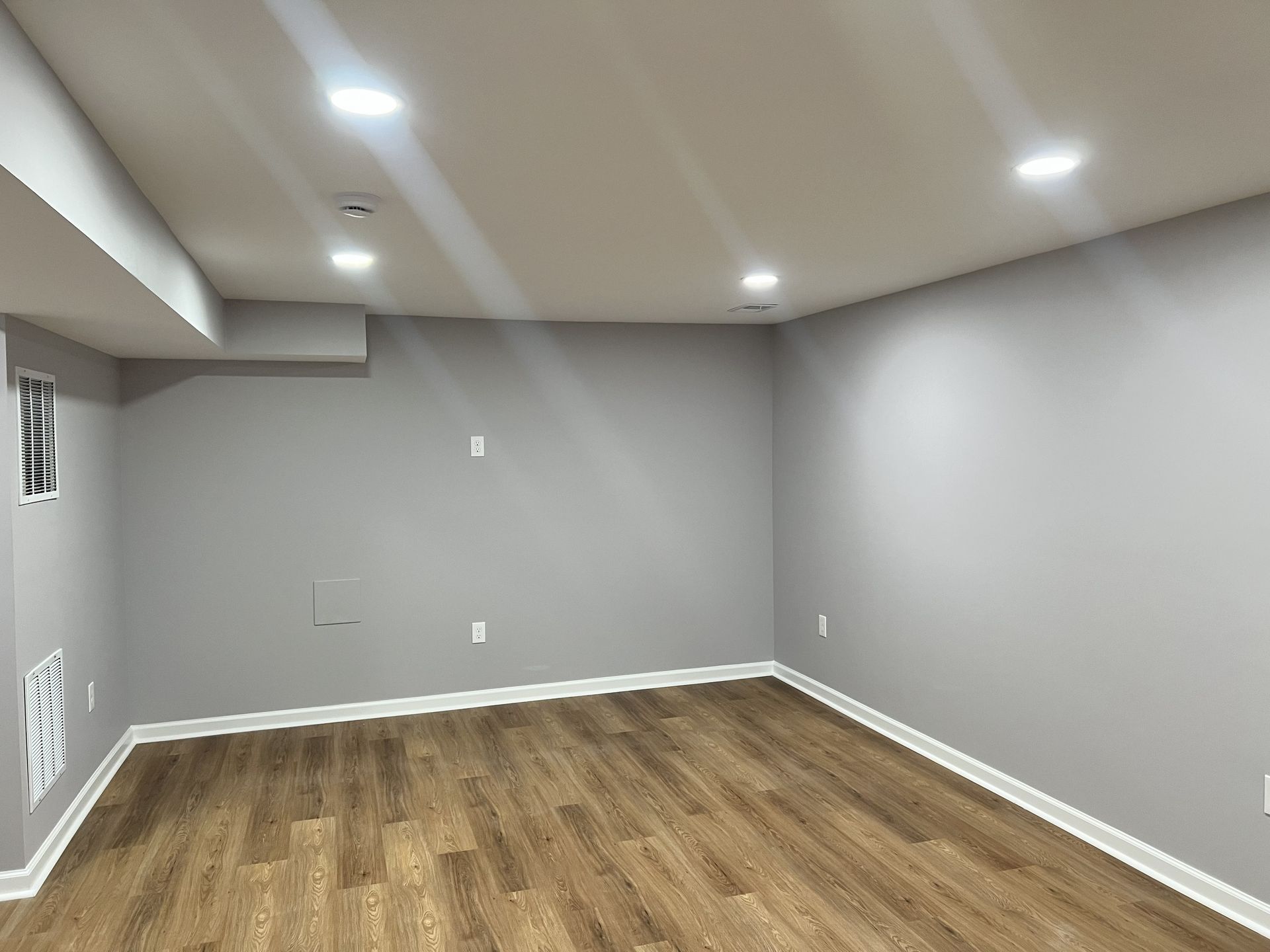 Empty gray basement room with wood flooring, recessed lights, and small basement windows