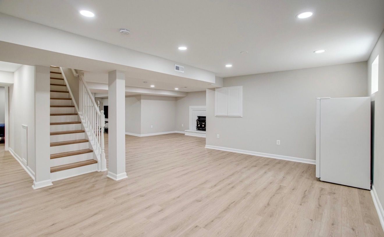 Bright empty basement room with light wood floors, white walls, stairs, and recessed lighting