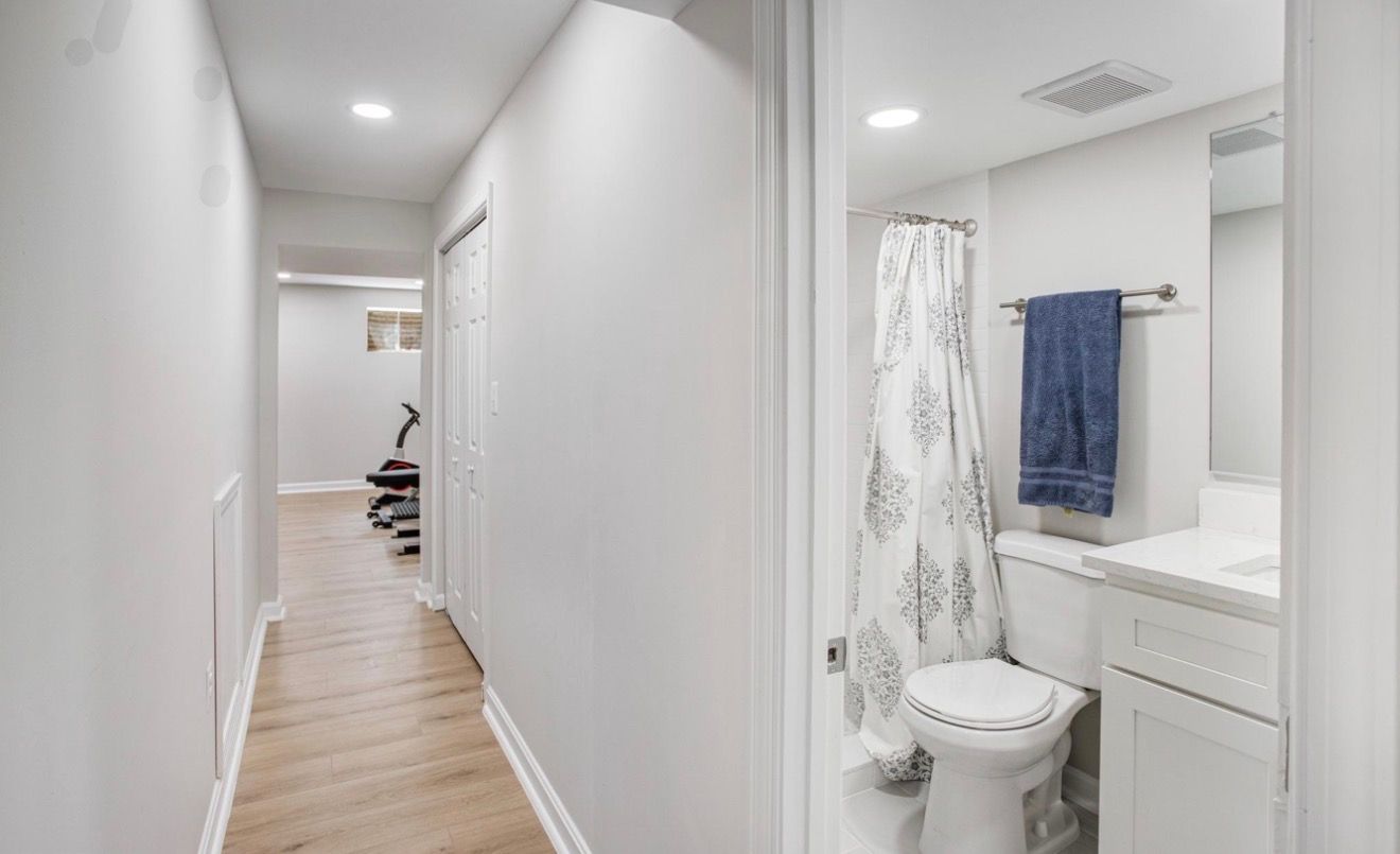 Bright hallway beside a small bathroom with white walls, a sink, toilet, towel, and shower curtain.