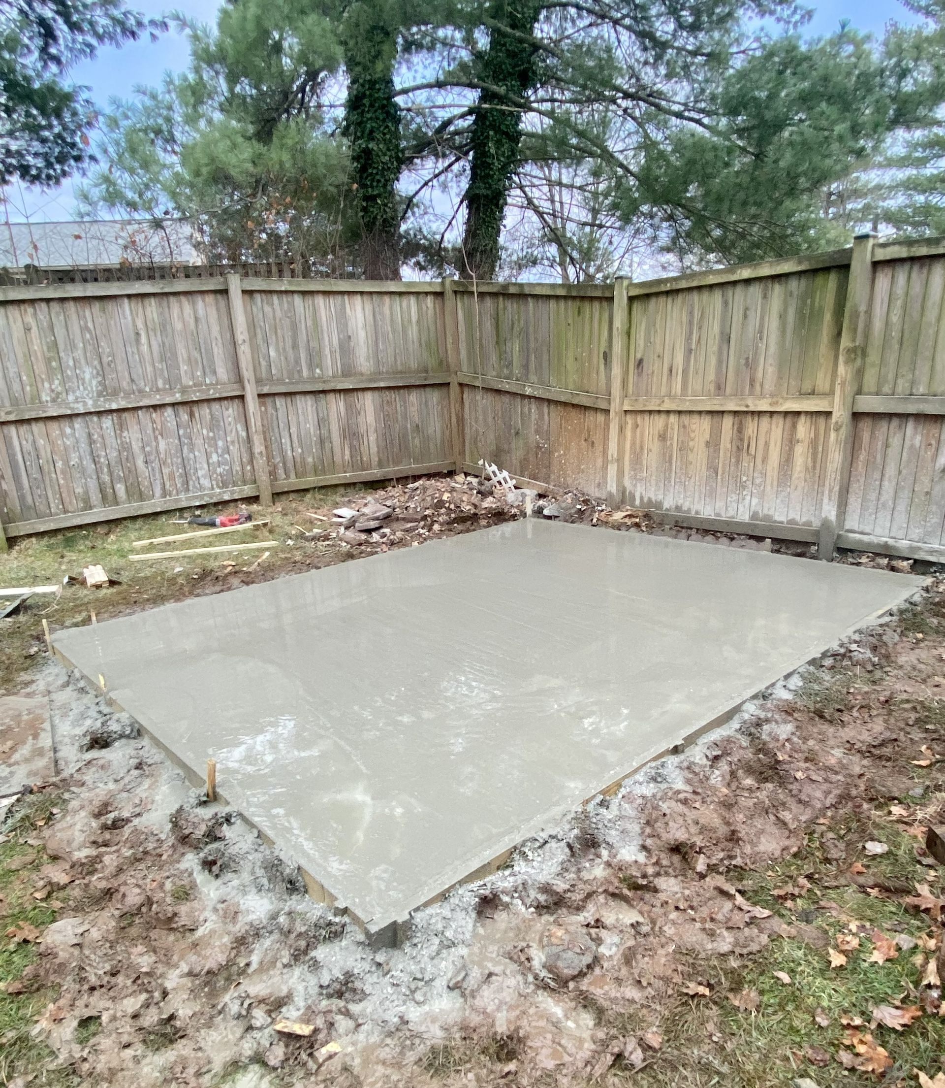 Fresh concrete slab in a fenced backyard corner with trees and leaf-strewn ground around it