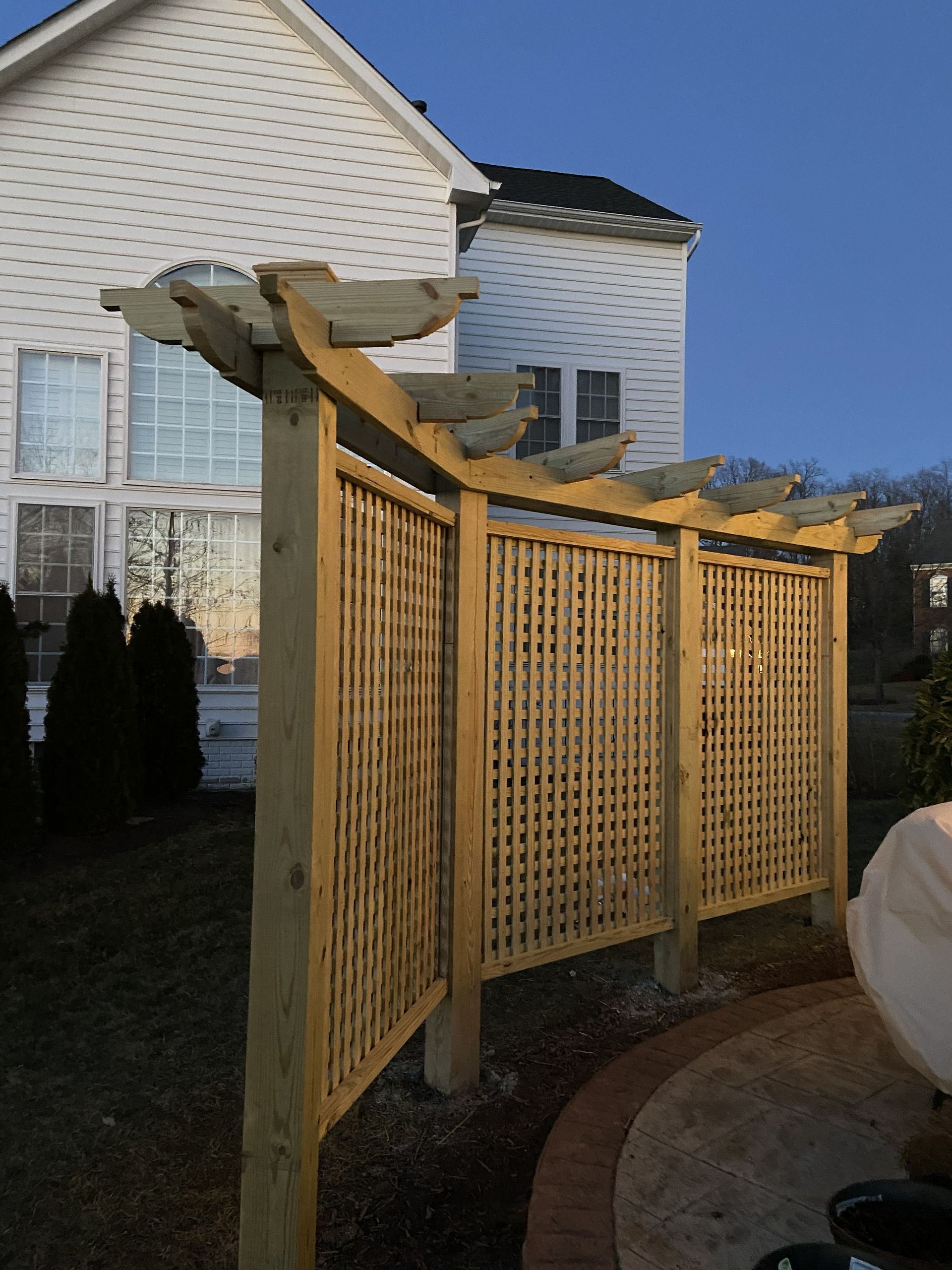 Wooden lattice pergola beside a white house in a backyard, with a curved stone patio and blue sky.