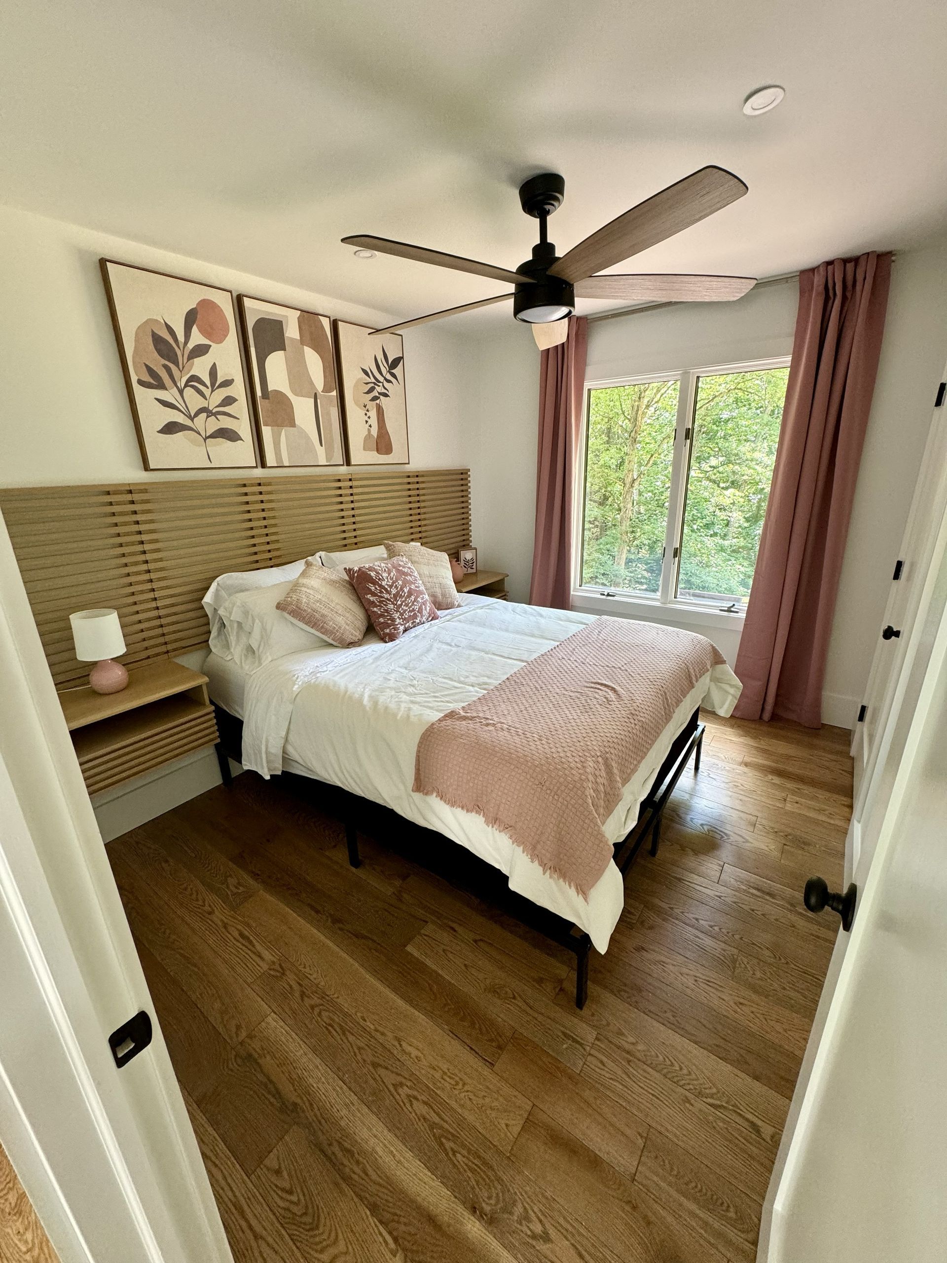 Bright bedroom with a bed, ceiling fan, floral wall art, pink curtains, and a window overlooking greenery