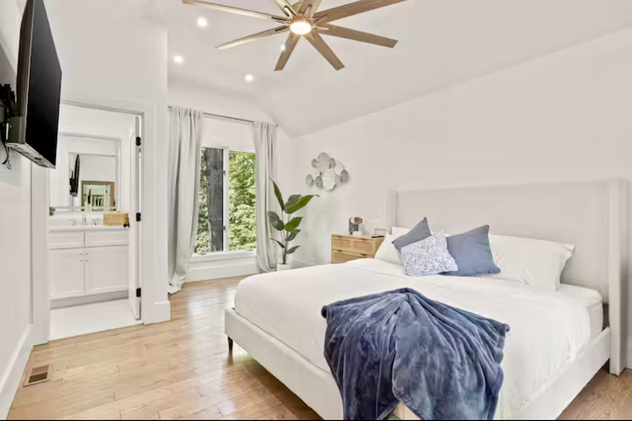 Bright bedroom with a white bed, blue throw blanket, ceiling fan, and window with greenery outside